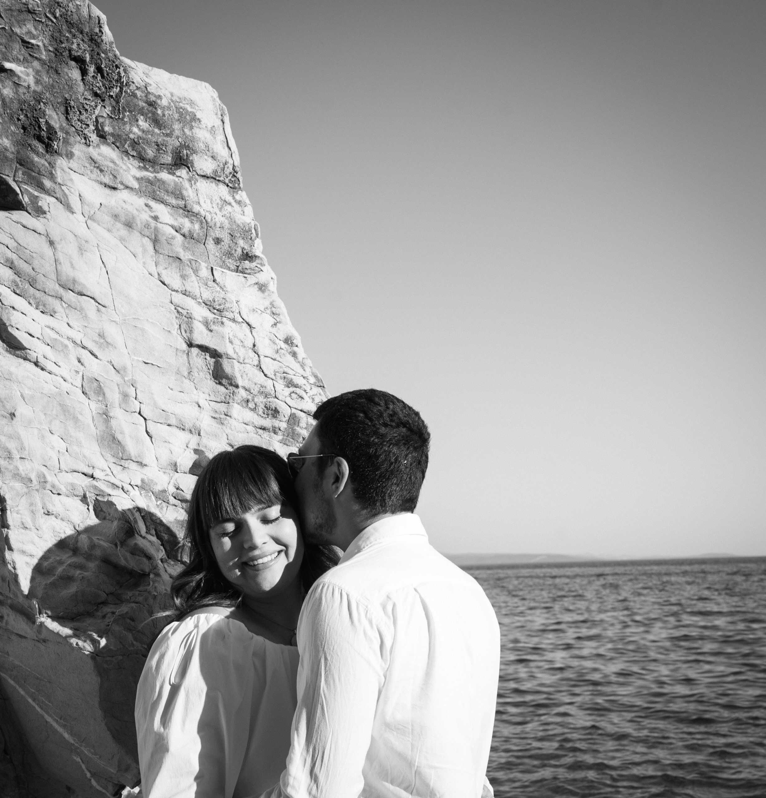 Zaruke na Obojenoj u Splitu; zaručnik ljubi mladenku u obraz dok su naslonjeni na visoku stijenu iznad plićaka.