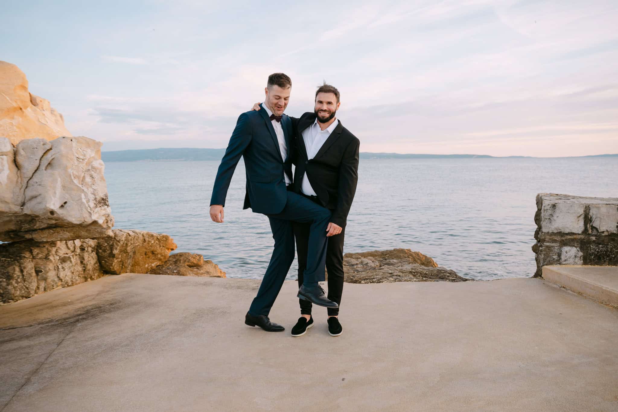 Mladenac Stipe i kum poziraju uz more na plaži Bačvice; opuštena fotografija u odijelima pod kamenitom stijenom u Splitu.