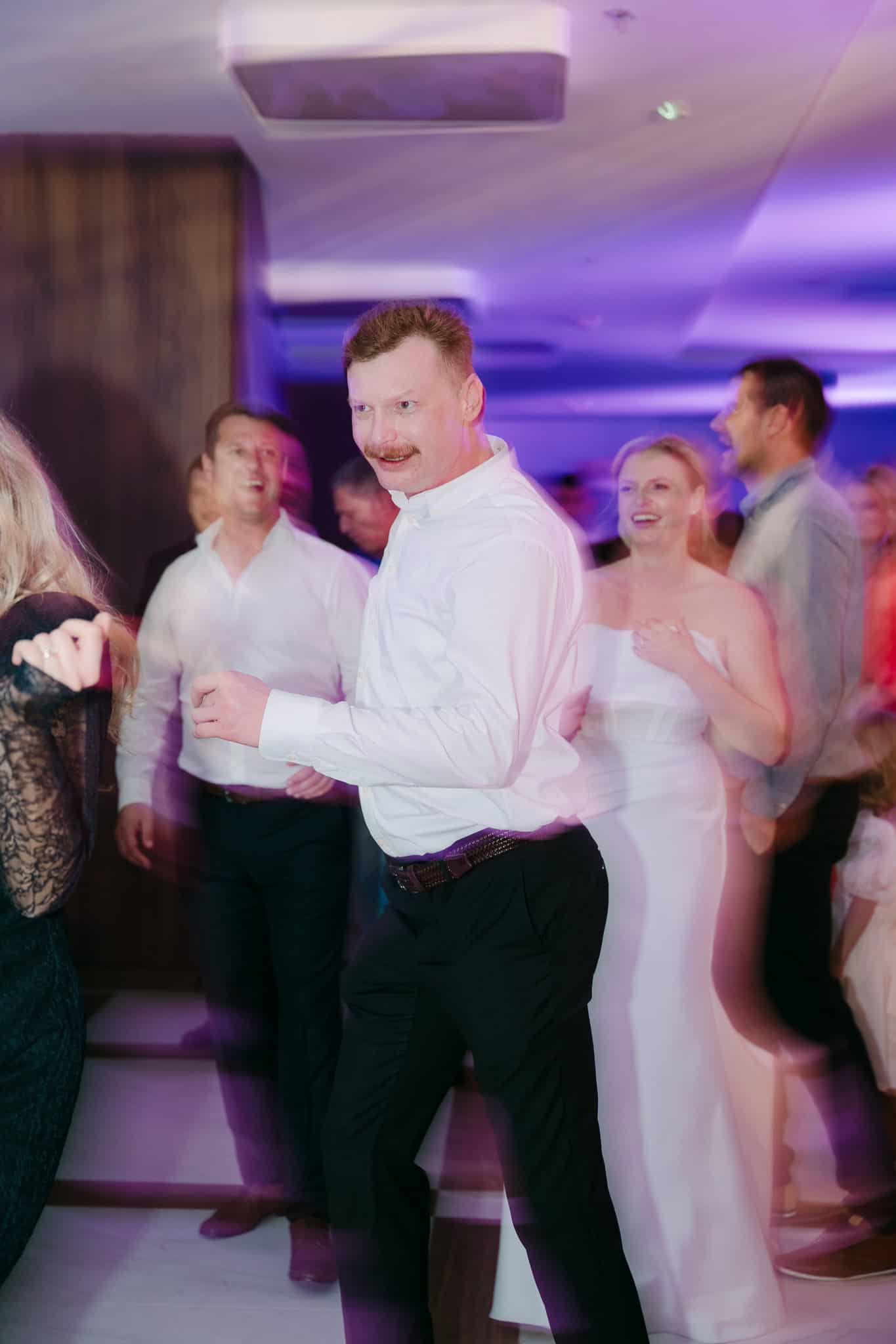 Energija i nezaustavljiv pokret na podiju za ples; nasmijani uzvanik u bijeloj košulji prepušta se ritmu, uz mladenku u pozadini.