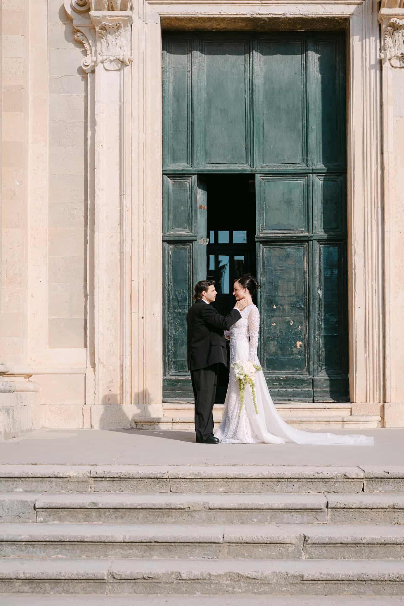 Dubrovnik session na otvorenom – nježan portret mladenaca; par u romantičnom trenutku stoji na kamenim stepenicama ispred povijesnih, masivnih drvenih vrata i raskošne arhitekture.