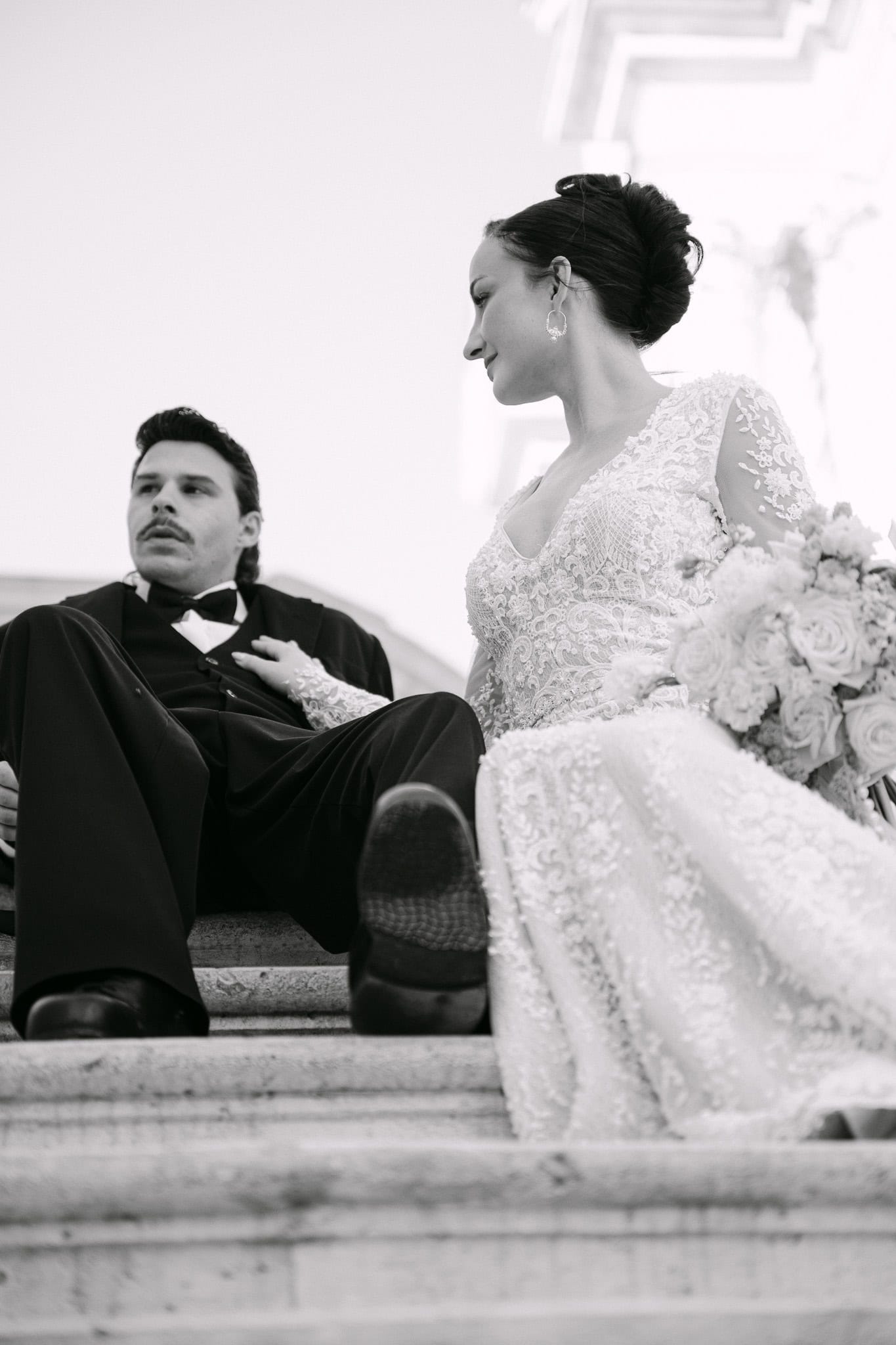 Filmski kadar para koji odmara na starinskom stubištu; mladenka nježno drži ruku na mladoženjinim prsima dok ga zaljubljeno gleda tijekom njihovog posebnog dana u Dubrovniku.