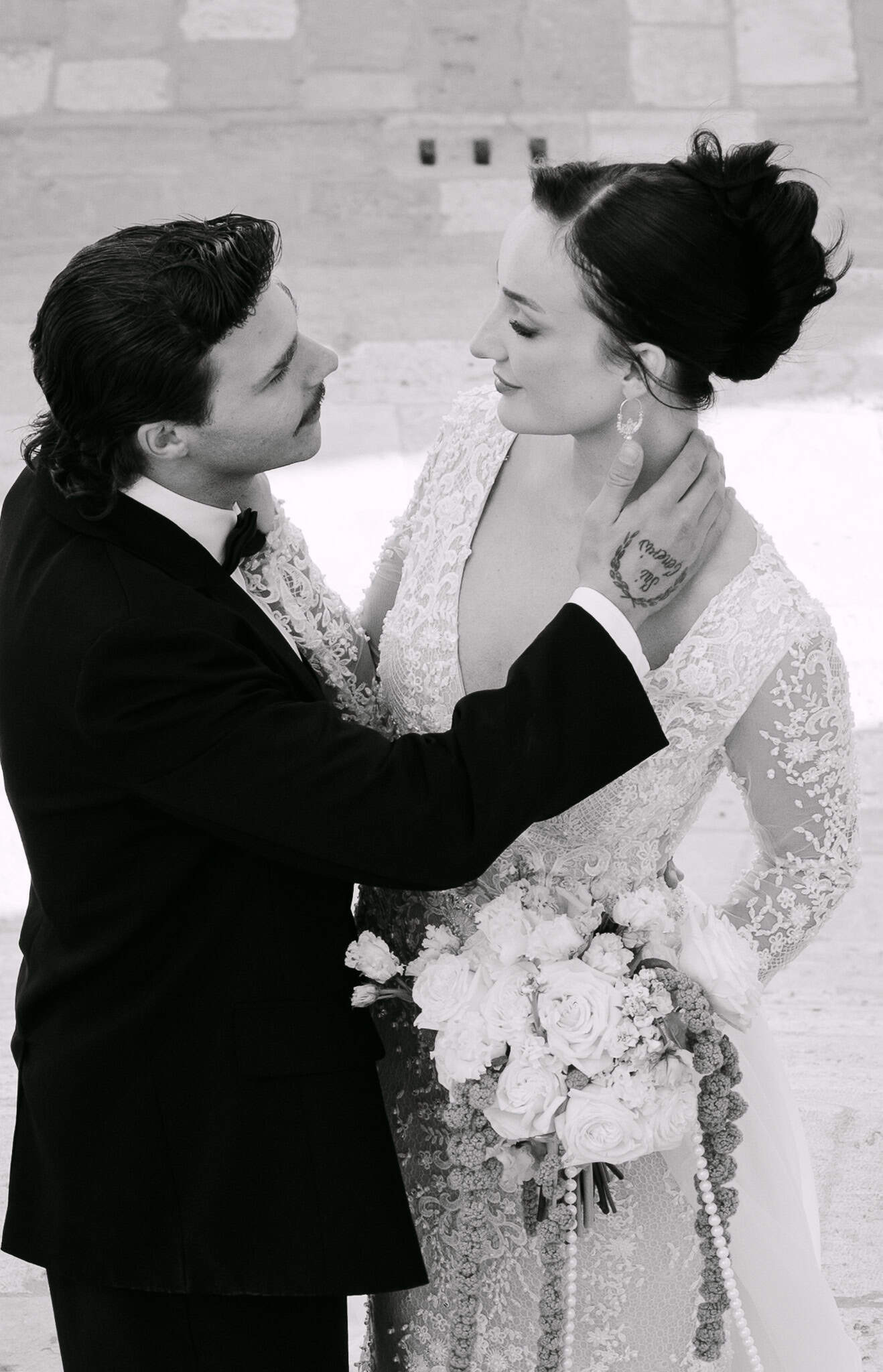 Mladoženja s vidljivom tetovažom na ruci nježno dodiruje mladenku ispred povijesnih zidova Dubrovnika.