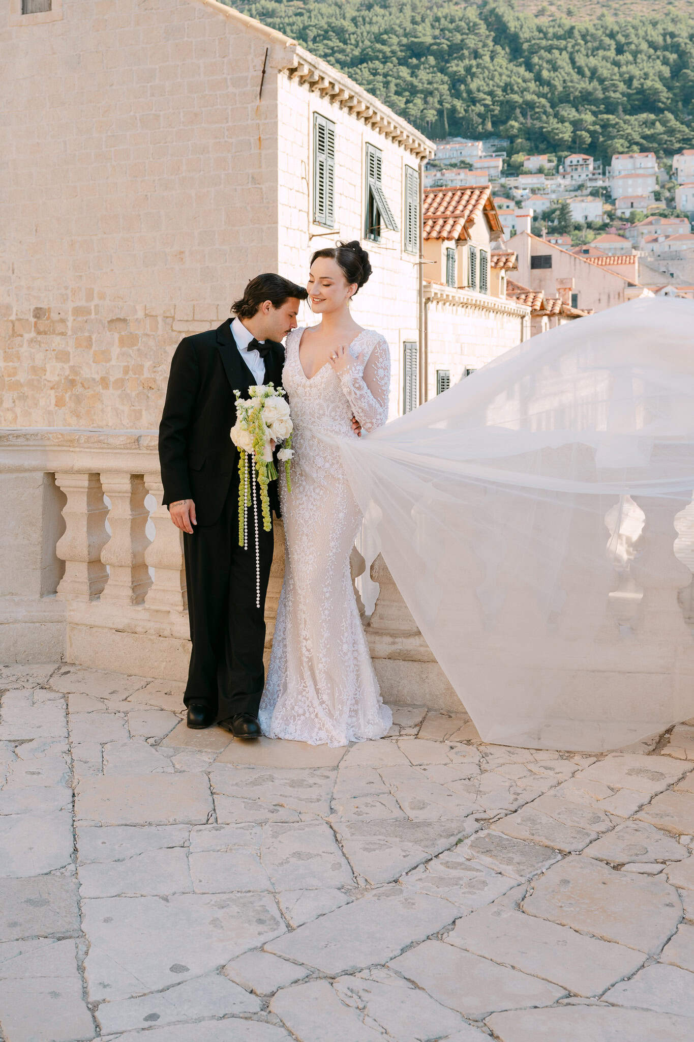 Nježni portret mladenaca na otvorenom; par u intimnom trenutku naslonjen na teksturirani kameni zid, držeći vjenčani buket.