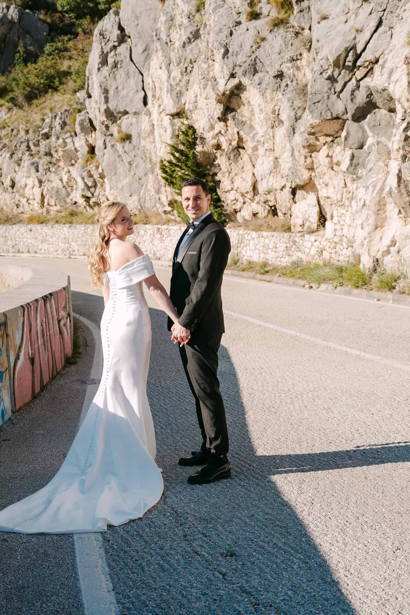 Nasmiješeni mladenci drže se za ruke na cesti ispod velike kamene litice tijekom svadbenog fotografiranja na otvorenom.