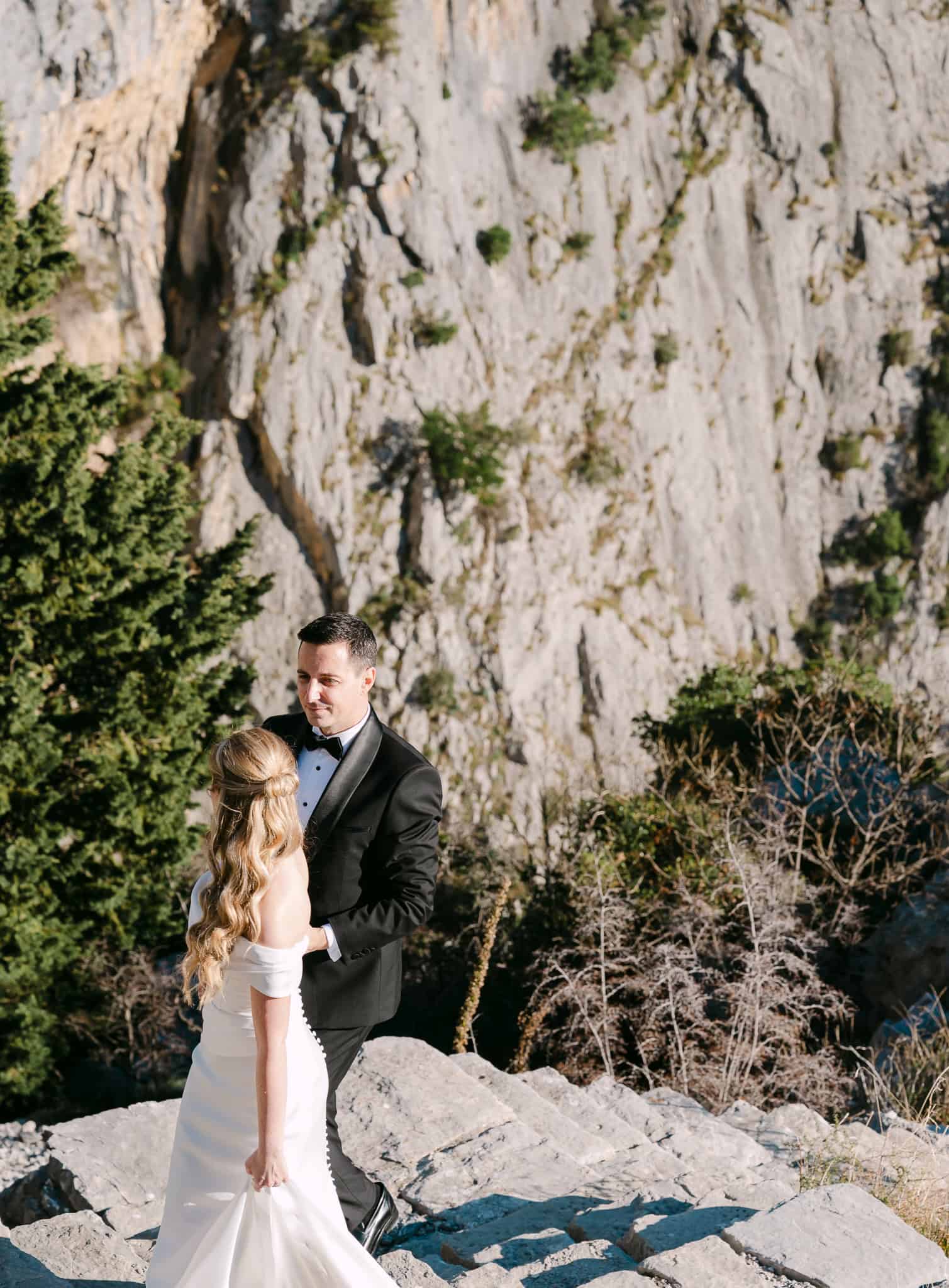 Mladenci se drže za ruke na kamenom usponu tijekom svadbenog fotografiranja u prirodi okruženi visokim stijenama.
