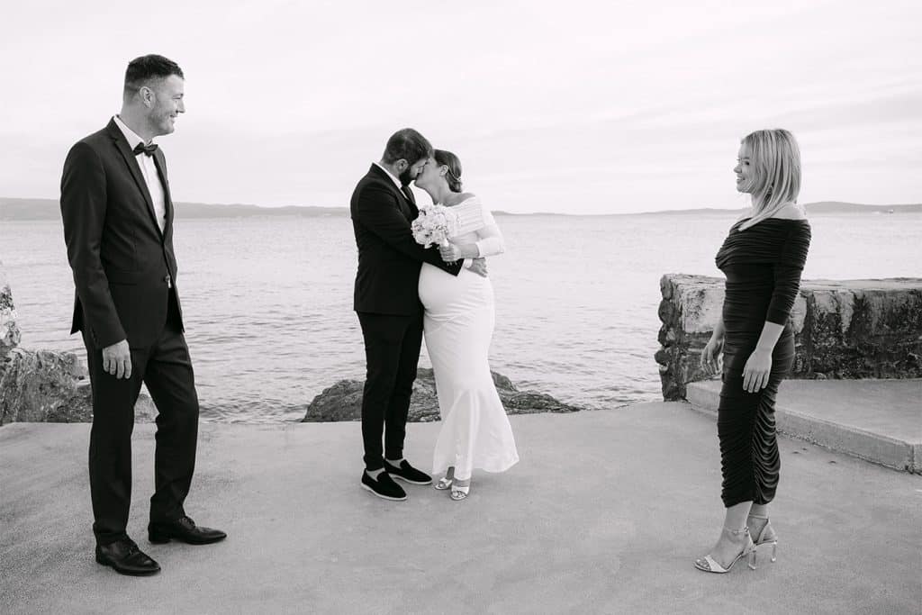 Mladenci u vjenčanici i odijelu s kumovima poziraju na kamenoj obali uz more tijekom svadbenog sessiona u Splitu.