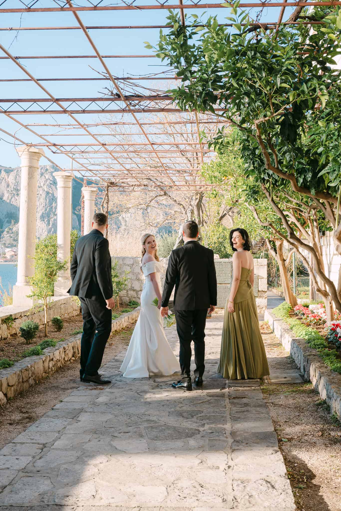 Mladenci s kumovima šetaju kamenom stazom uz more ispod pergole tijekom fotografiranja vjenčanja u Omiš.