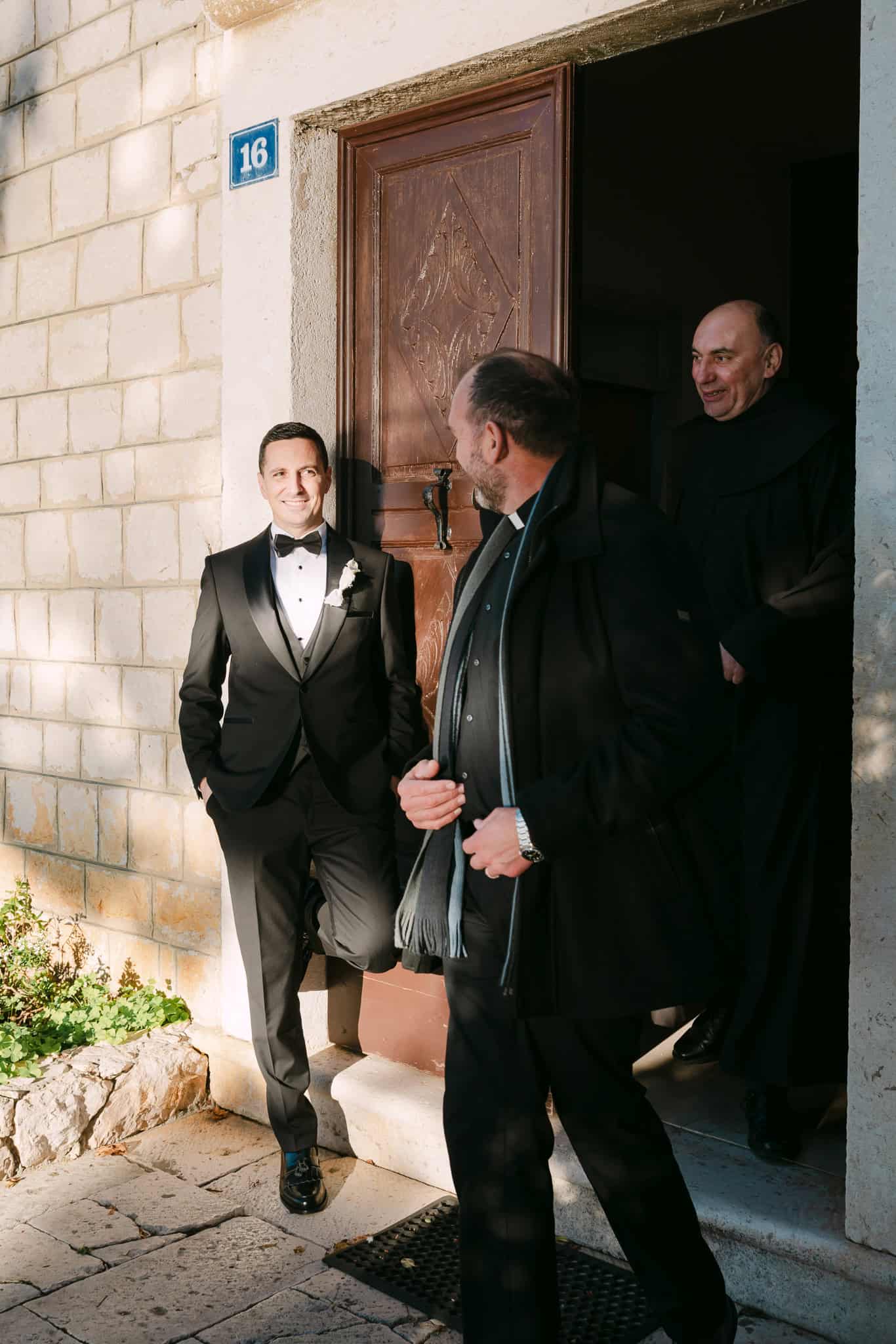 Mladoženja stoji ispred kamenih vrata crkve u Omišu u razgovoru sa svećenikom prije vjenčanja.