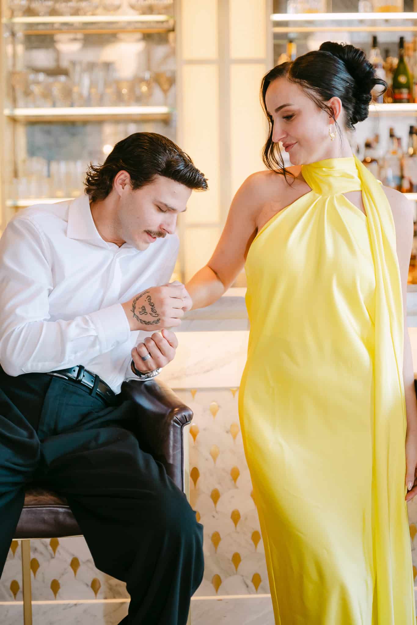 Dubrovnik, vjenčani session - nježan trenutak uz mramorni šank; mladenka u elegantnoj žutoj haljini gleda u mladoženjinu ruku s tetovažom.