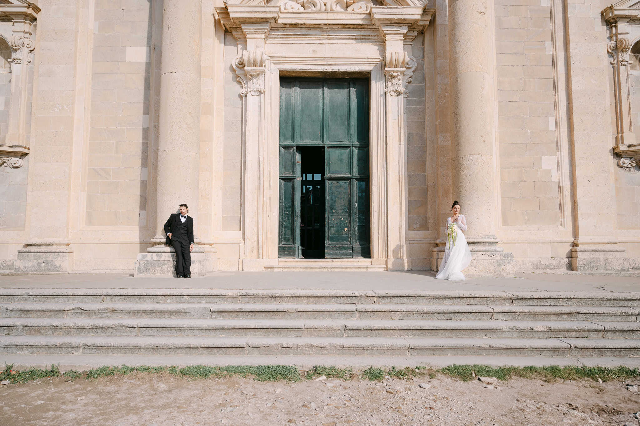 Moderan lifestyle portret; mladoženja i mladenka ležerno poziraju svatko uz svoj kameni stup ispred velikih starinskih vrata i suncem okupanih zidina u gradu Dubrovniku.