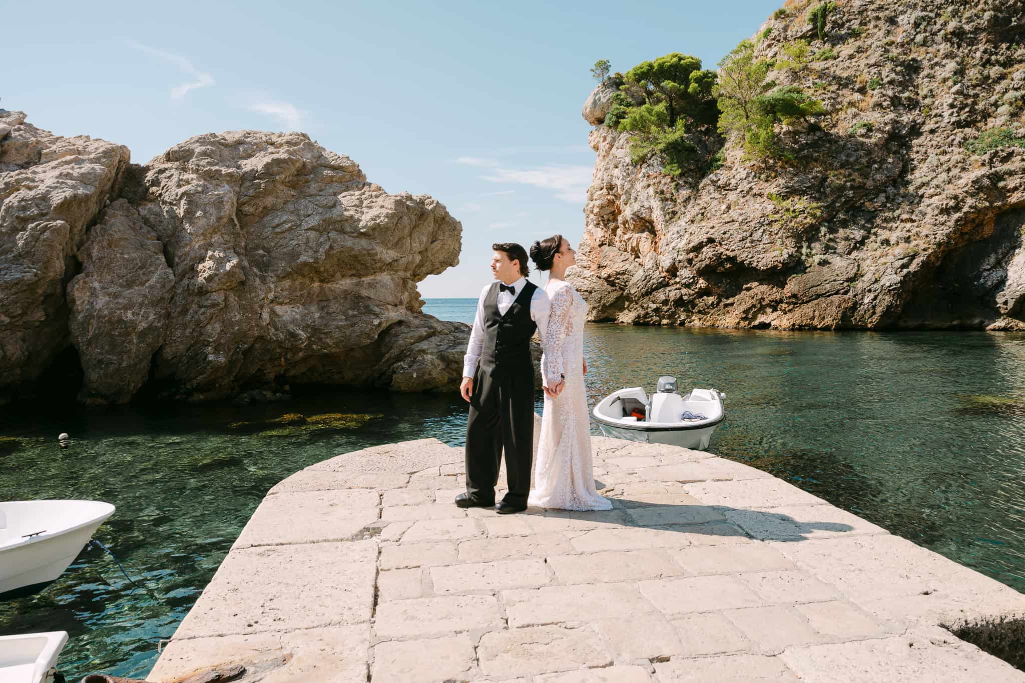 Dubrovnik destination wedding - moderan editorijalni portret na obali; mladenci u stiliziranoj pozi na sunčanom mulu ispred mora i visokih stijena.