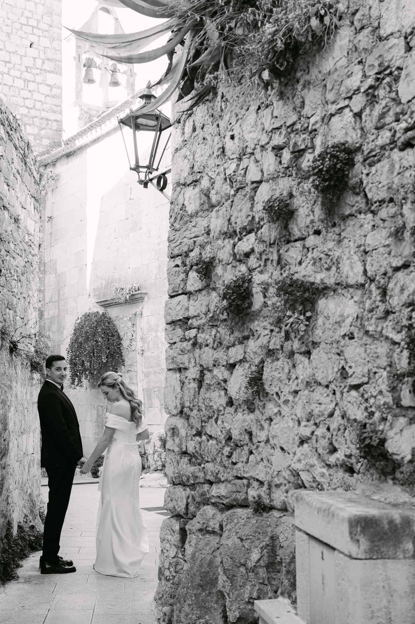 Crno-bijela fotografija mladenaca koji šeću kamenom ulicom starog grada s pogledom preko ramena prema objektivu, uz crkveni zvonik u pozadini.