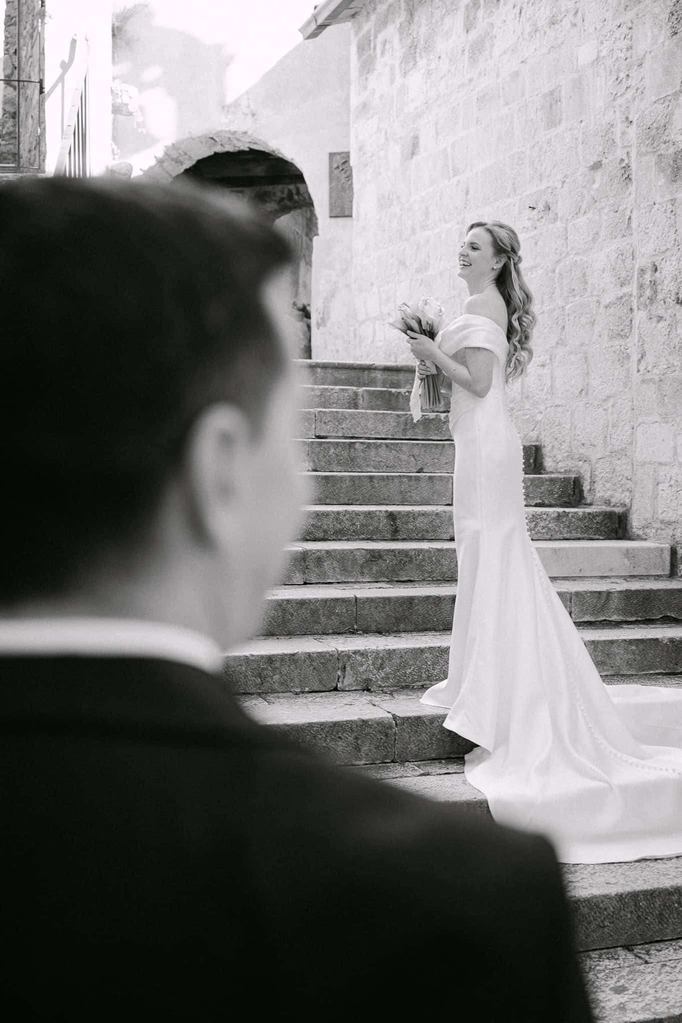 Crno-bijeli kadar mladenke u vjenčanici koja stoji na vanjskim kamenim stepenicama i nasmijano gleda unatrag prema mladoženji u prednjem planu.