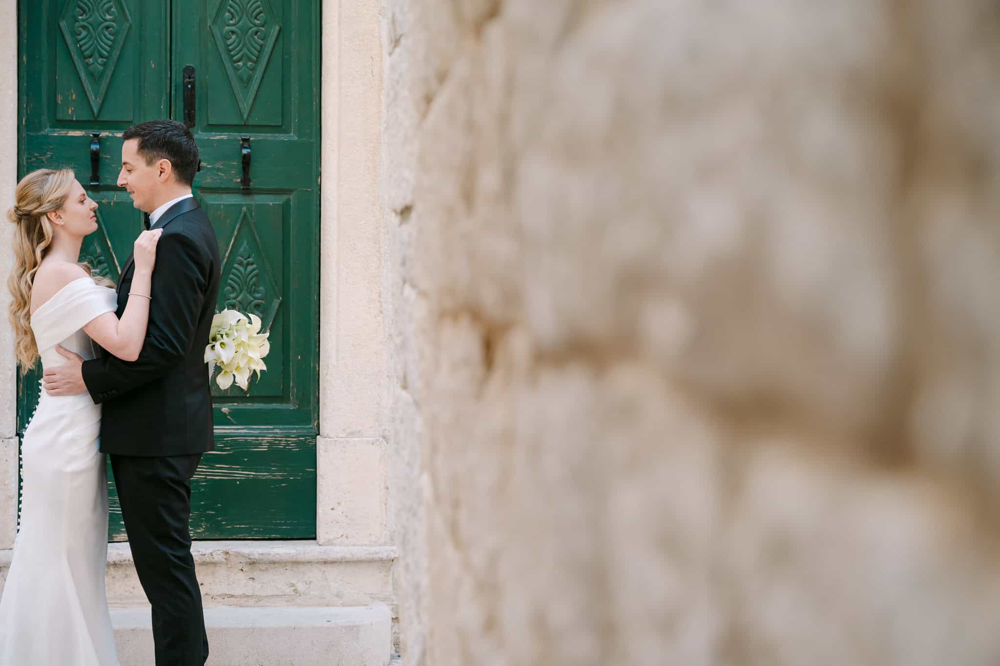 Mladenci stoje zagrljeni ispred starih masivnih zelenih vrata na kamenoj kući tijekom fotografiranja vjenčanja u Dalmaciji.