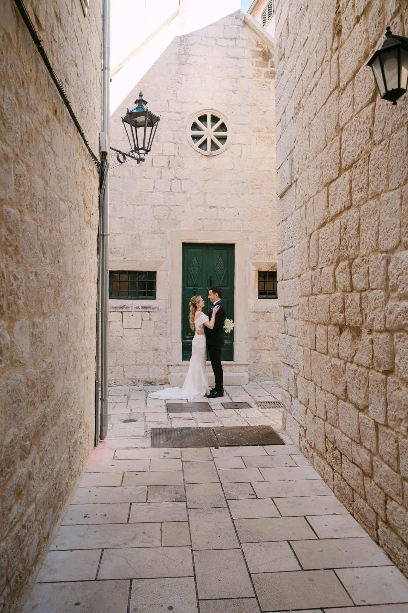 Široki kadar mladenaca koji stoje u uskoj dalmatinskoj kamenoj ulici grada Omiša ispred zelenih vrata i zidne ulične svjetiljke.