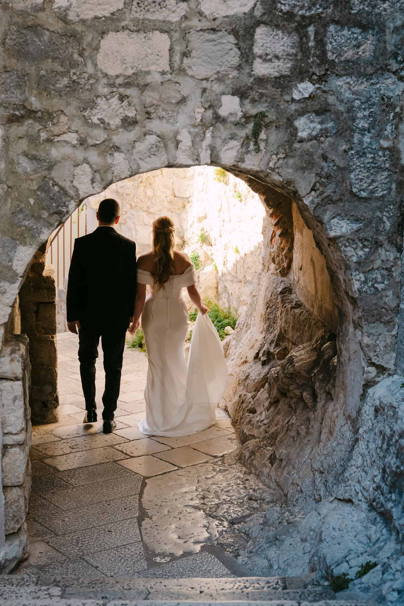 Mladenci se drže za ruke i prolaze ispod starog kamenog luka u povijesnom centru grada Omiša tijekom svadbenog fotografiranja.
