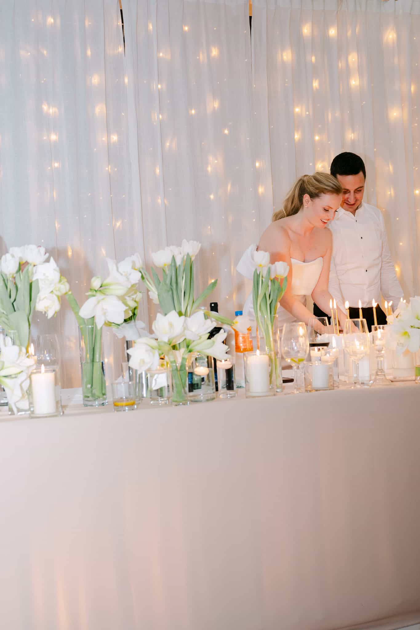 Svečani trenutak uz vjenčanu tortu; mladenci nagnuti jedno prema drugome pažljivo pale svjećice na torti okruženoj bogatim aranžmanima bijelih tulipana, obasjani toplom svjetlošću lampica u pozadini.