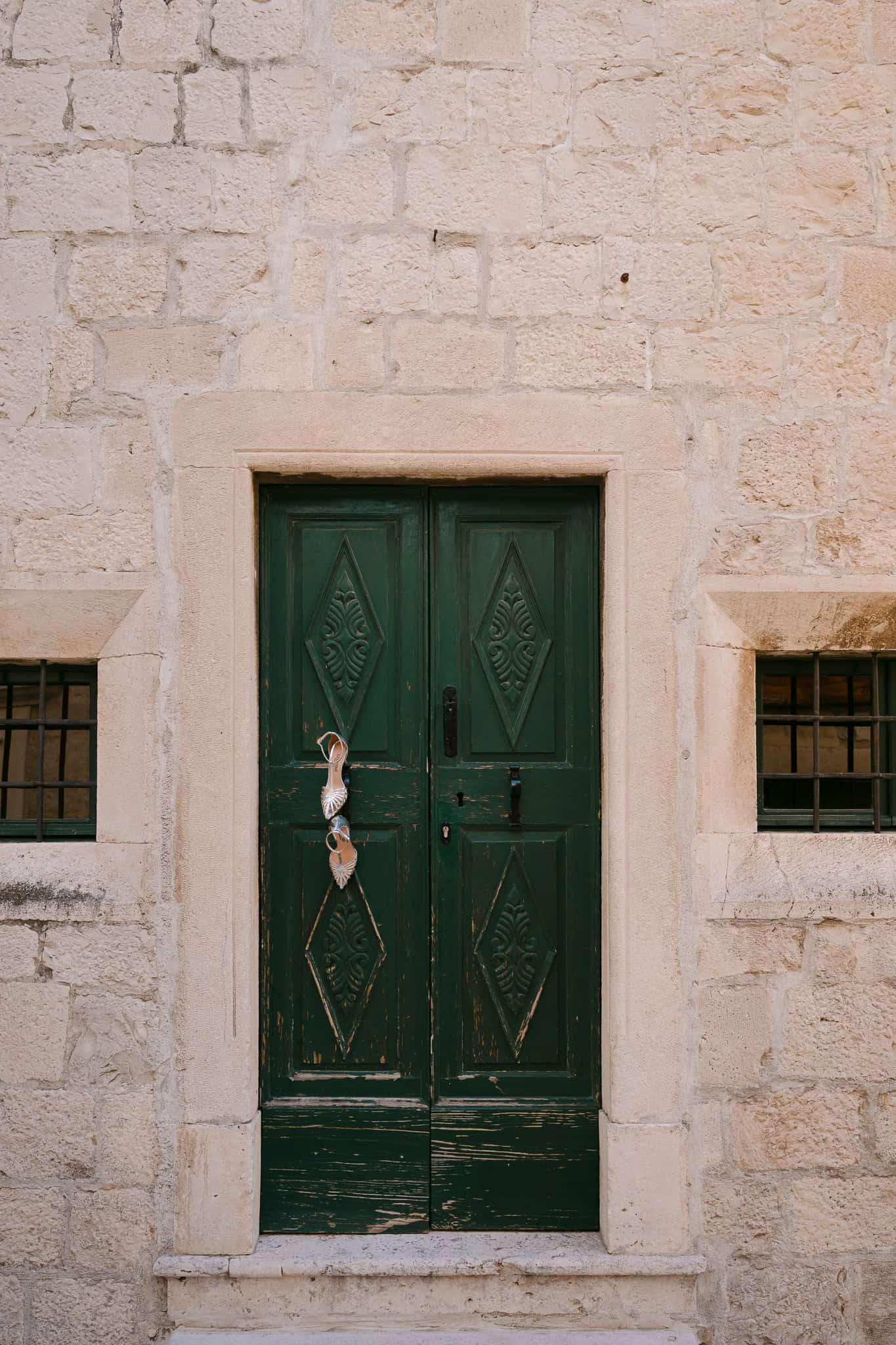 Vjenčane cipele na visoku petu vise na starim zelenim drvenim vratima dalmatinske kamene kuće tijekom priprema za vjenčanje.