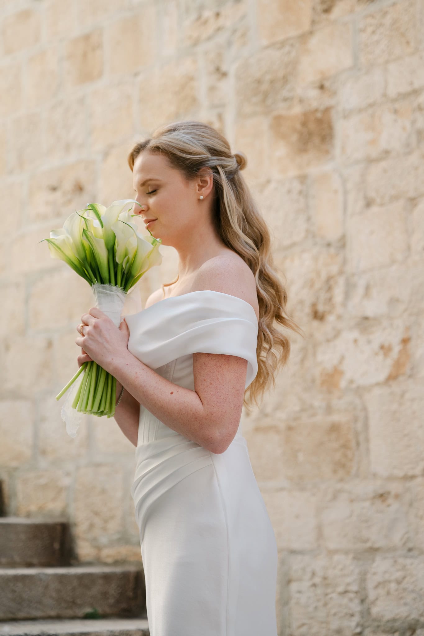 Mladenka u vjenčanici spuštenih ramena stoji ispred kamenog zida i miriše svadbeni bidermajer od bijelih kala.
