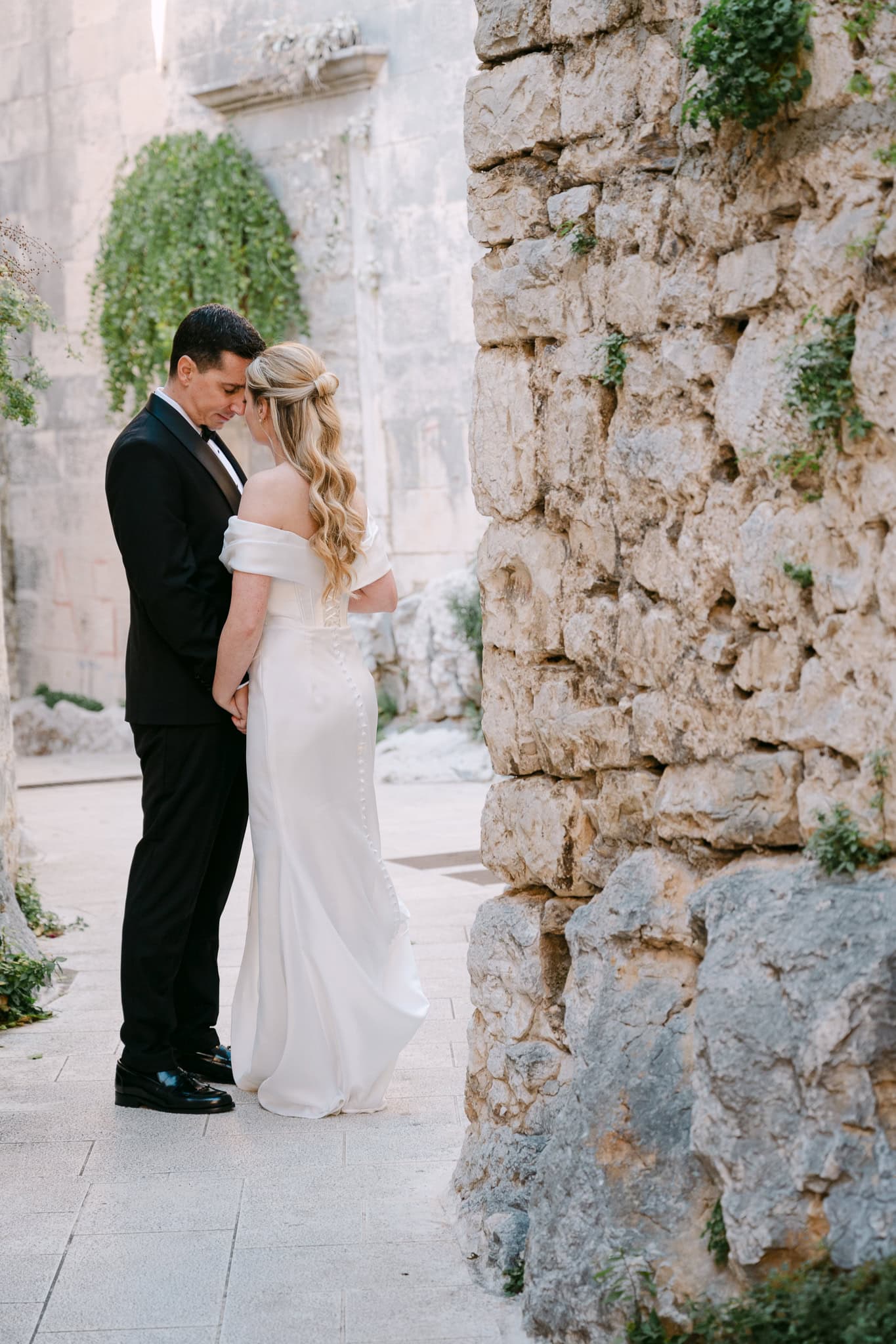 Mladenci naslonjeni glava na glavu drže se za ruke stojeći uz stari kameni zid okružen mediteranskim zelenilom.