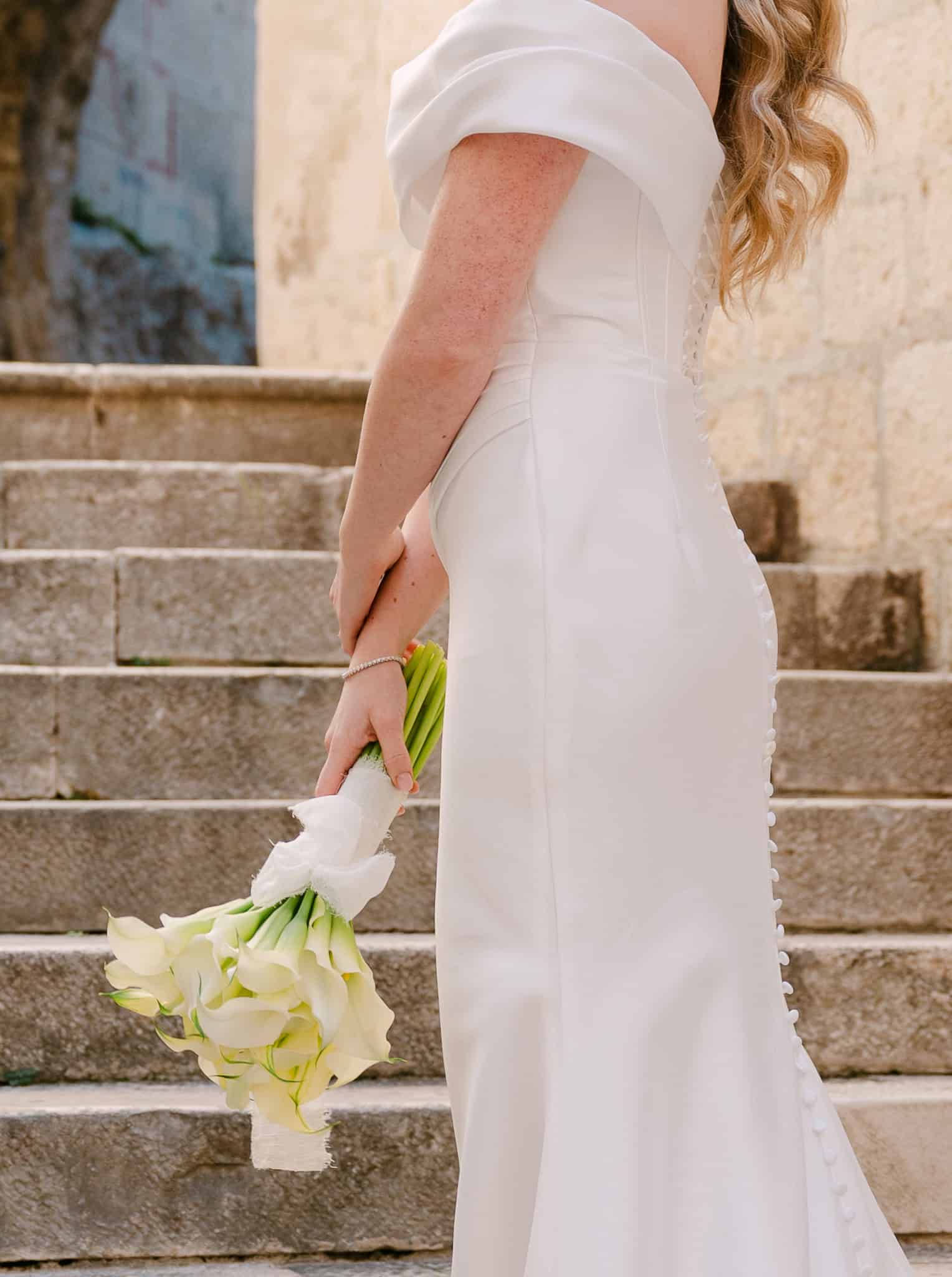 Detalj uske vjenčanice s nizom gumbića niz leđa i svadbenog buketa od bijelih kala kojeg mladenka drži uz tijelo na kamenim stepenicama.
