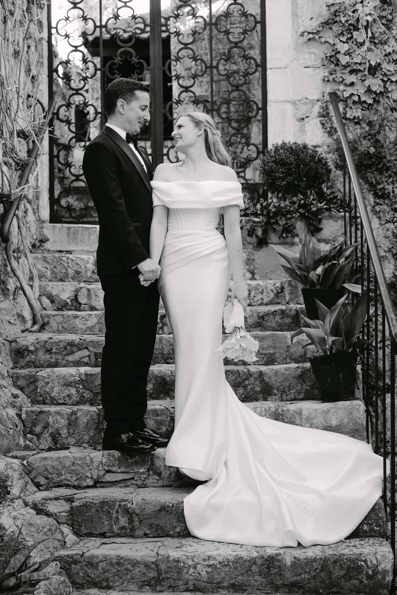 Crno-bijeli kadar mladenaca na vanjskom kamenom stubištu koji naglašava pad dugog šlepa vjenčanice niz stepenice.