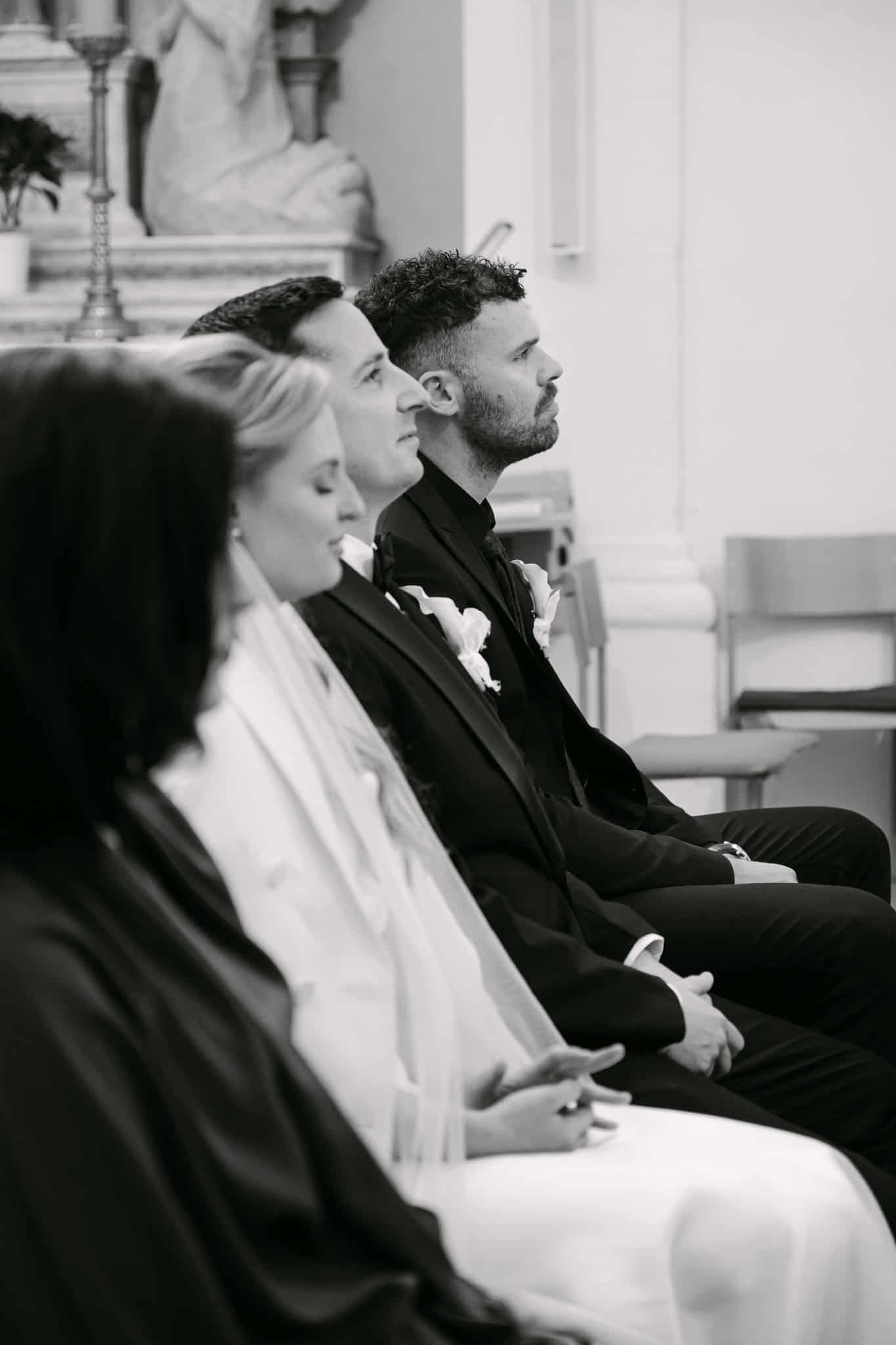 Crno-bijeli profilni kadar mladenaca i kumova koji sjede u crkvenim klupama i slušaju obred vjenčanja.