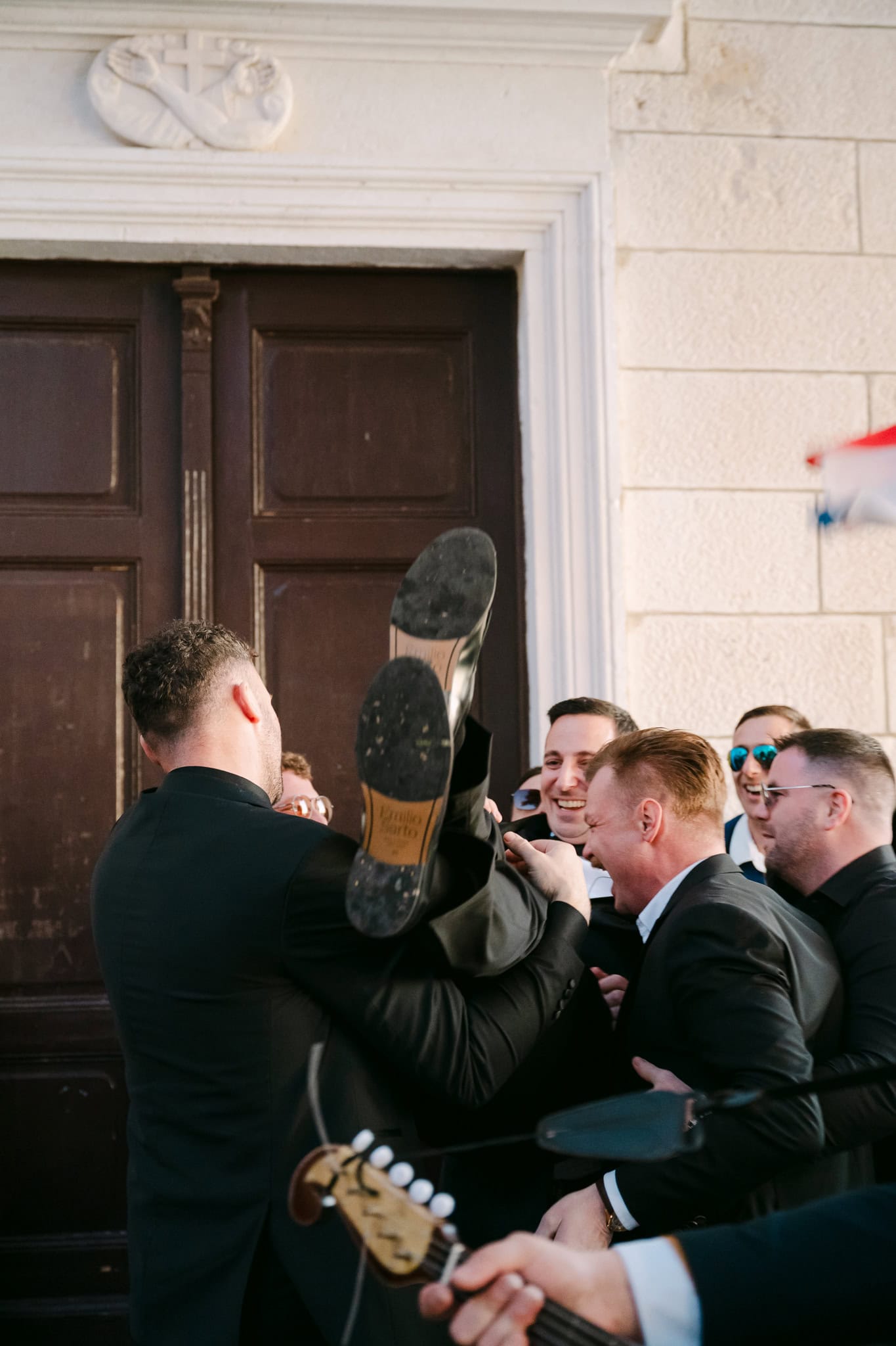 Prijatelji dižu mladoženju u zrak na vjenčanju ispred crkve, vesela atmosfera.