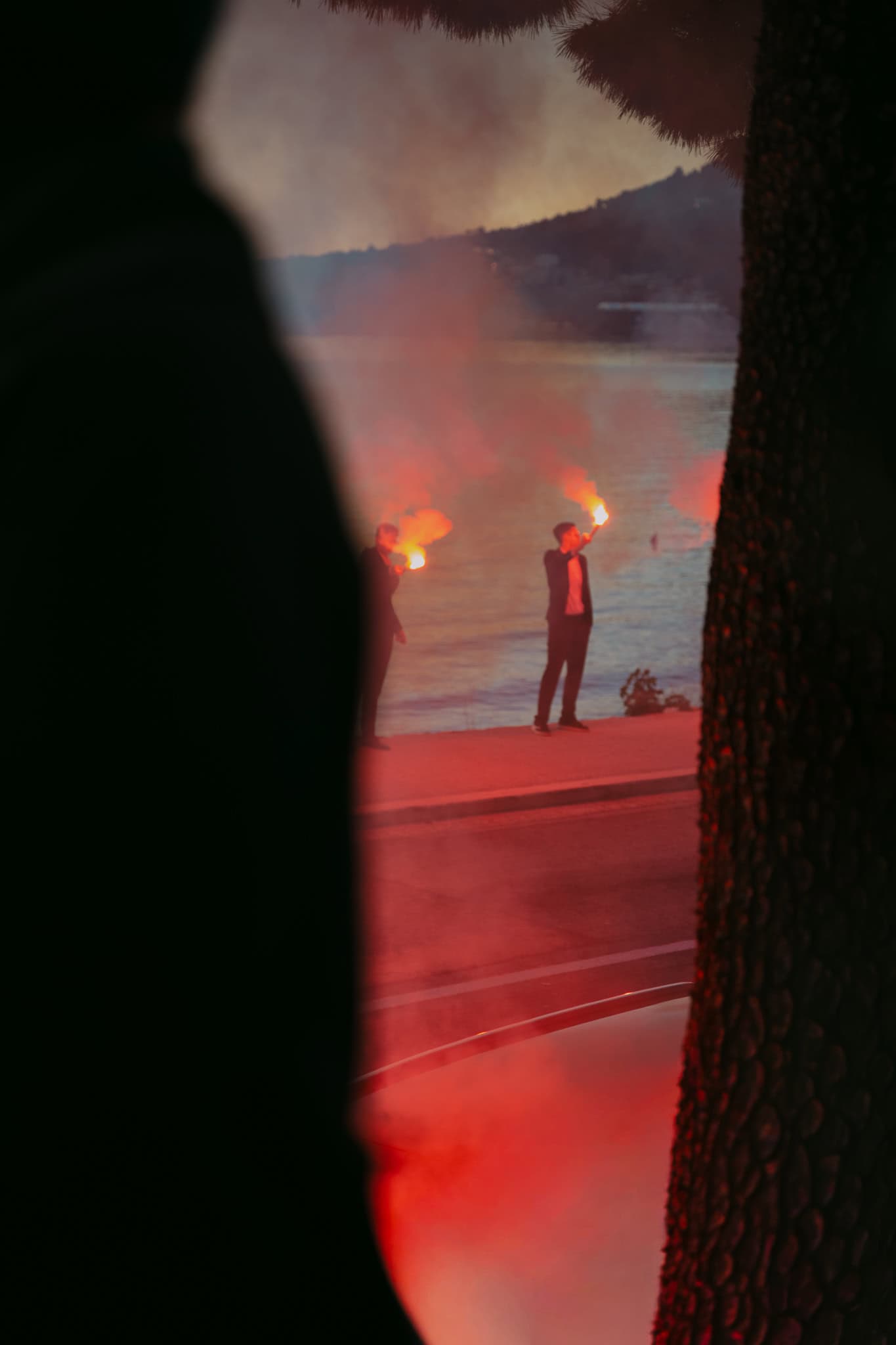 Svadbeno slavlje uz more gdje gosti drže upaljene baklje s crvenim dimom.