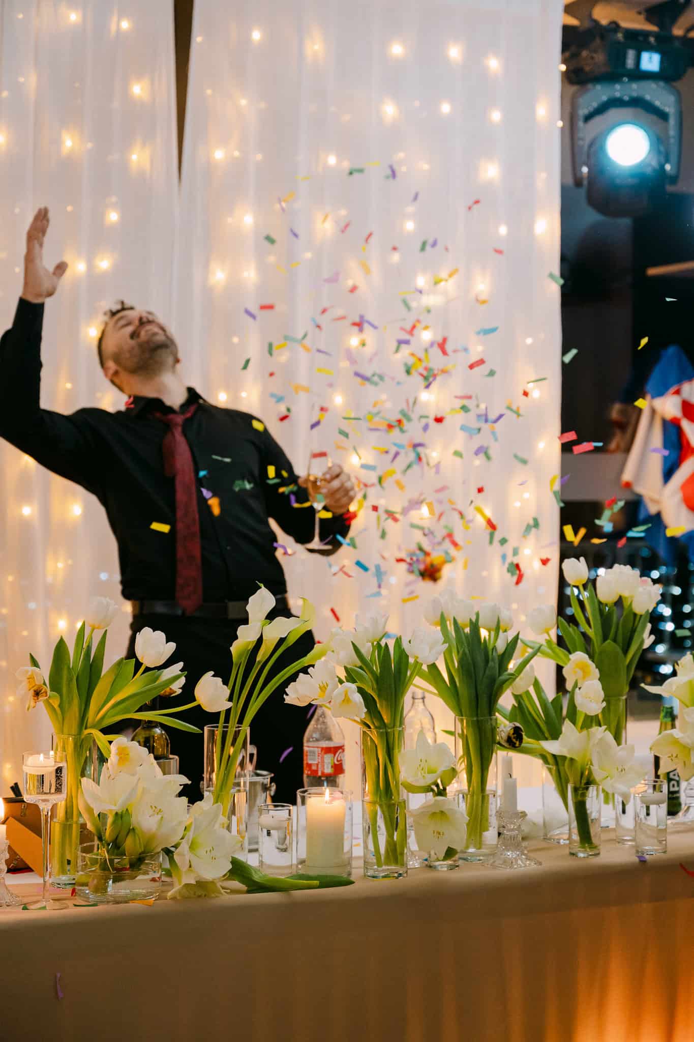 Spontani trenutak slavlja na svadbenoj večeri; raspoloženi mladoženjin kum uživa u plesu dok šareni konfeti padaju oko njega iza svečanog stola ukrašenog proljetnim bijelim tulipanima.