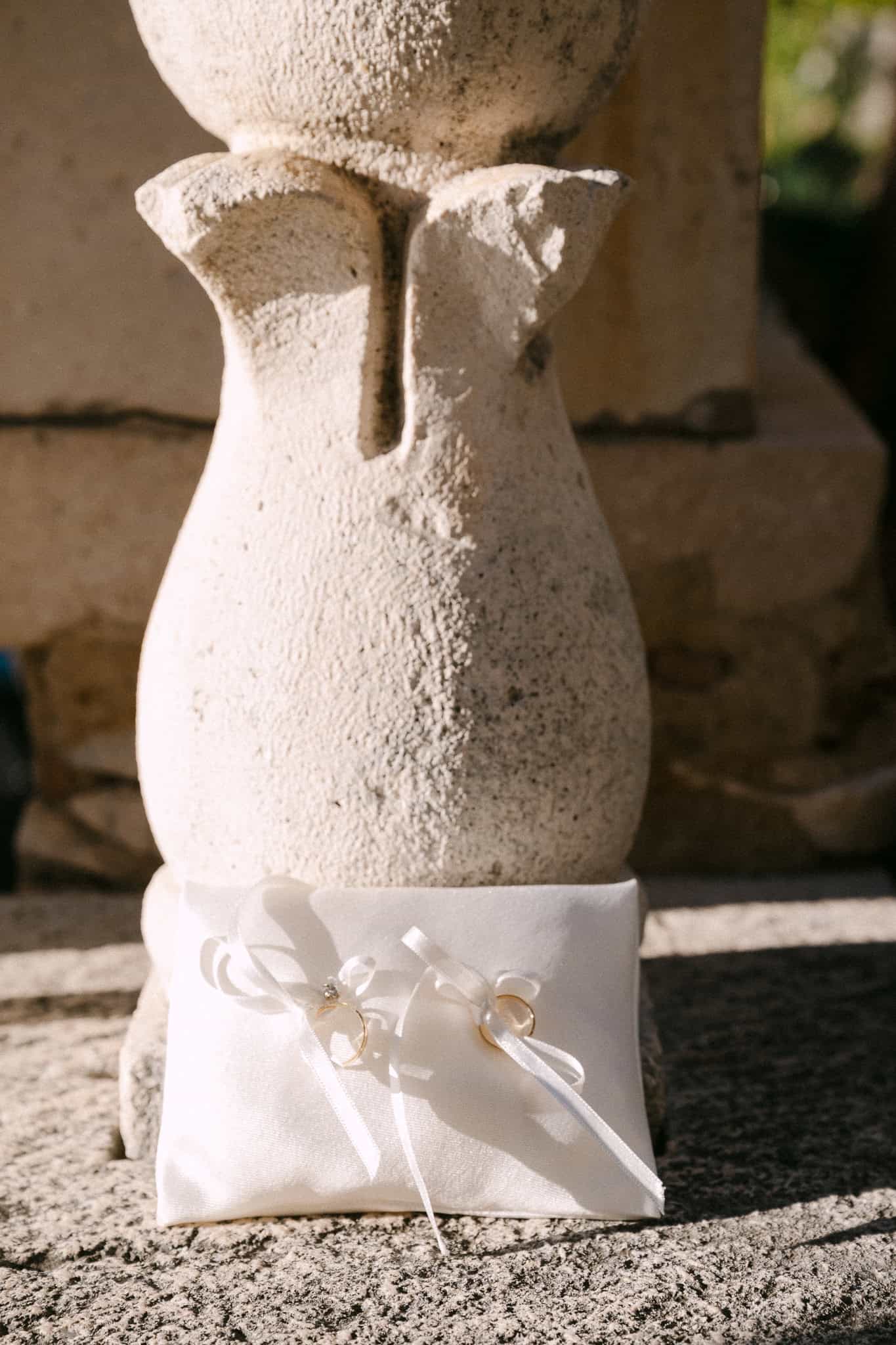 Vjenčano prstenje zavezano bijelim vrpcama za jastučić odloženo na kamenom stupu na otvorenom.