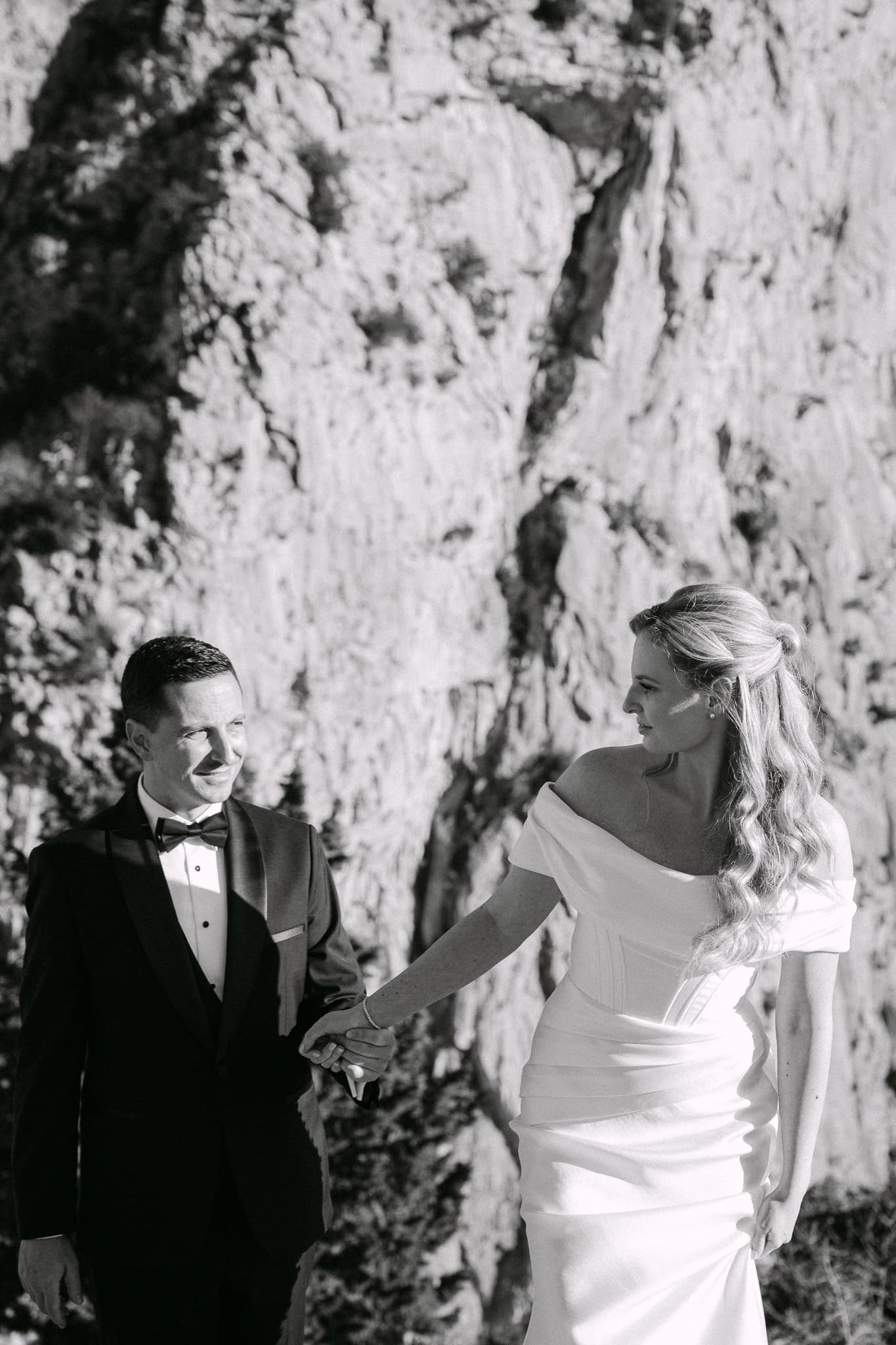 Mladenci se drže za ruke i hodaju ispred velike kamene litice na crno-bijeloj svadbenoj fotografiji.