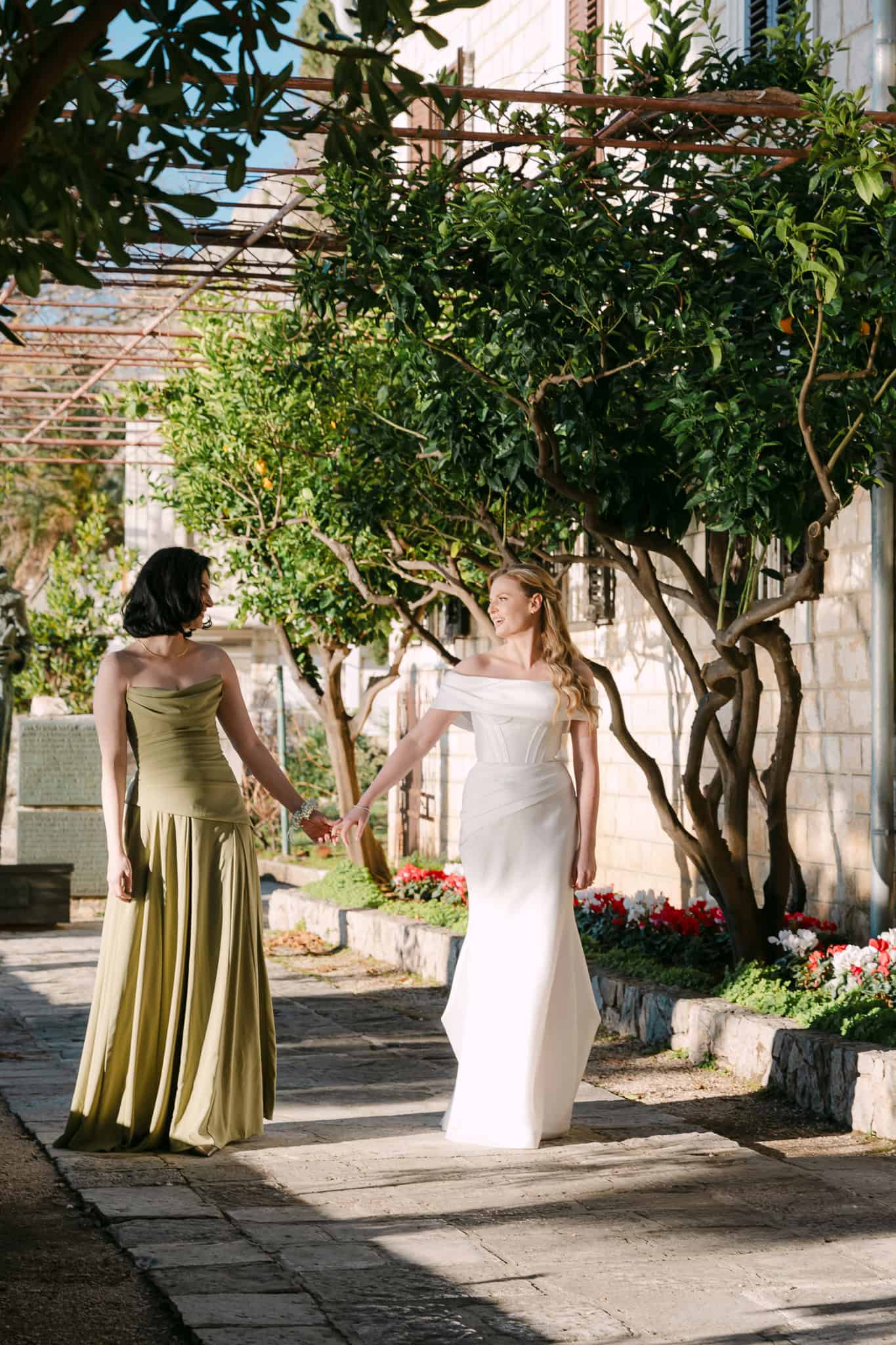 Mladenka i kuma u maslinastozelenoj haljini drže se za ruke i hodaju kamenom stazom ispod drveća u Omišu.