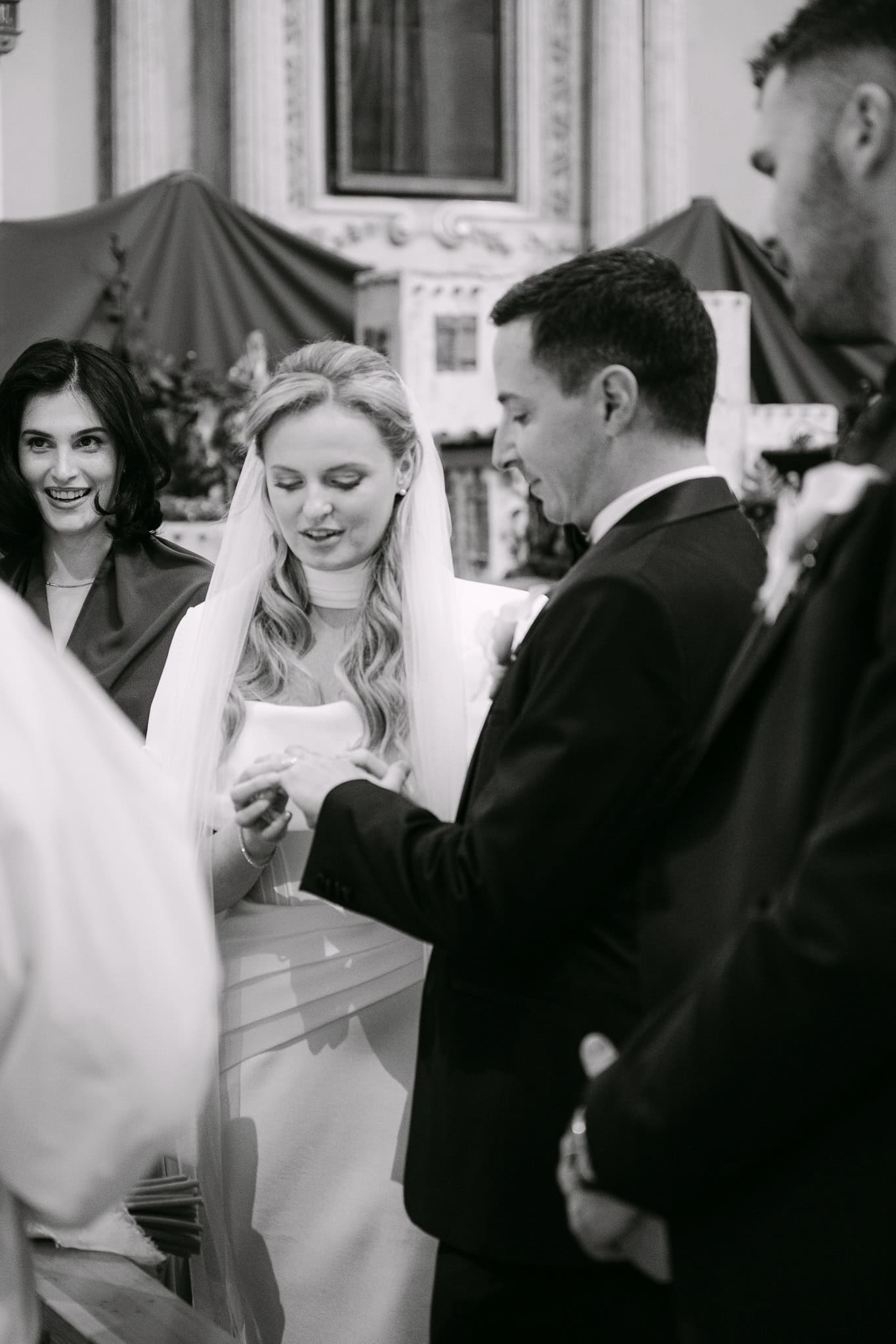 Crno-bijela fotografija obreda na kojoj mladenka stavlja vjenčani prsten na ruku mladoženje uz kumu u pozadini.