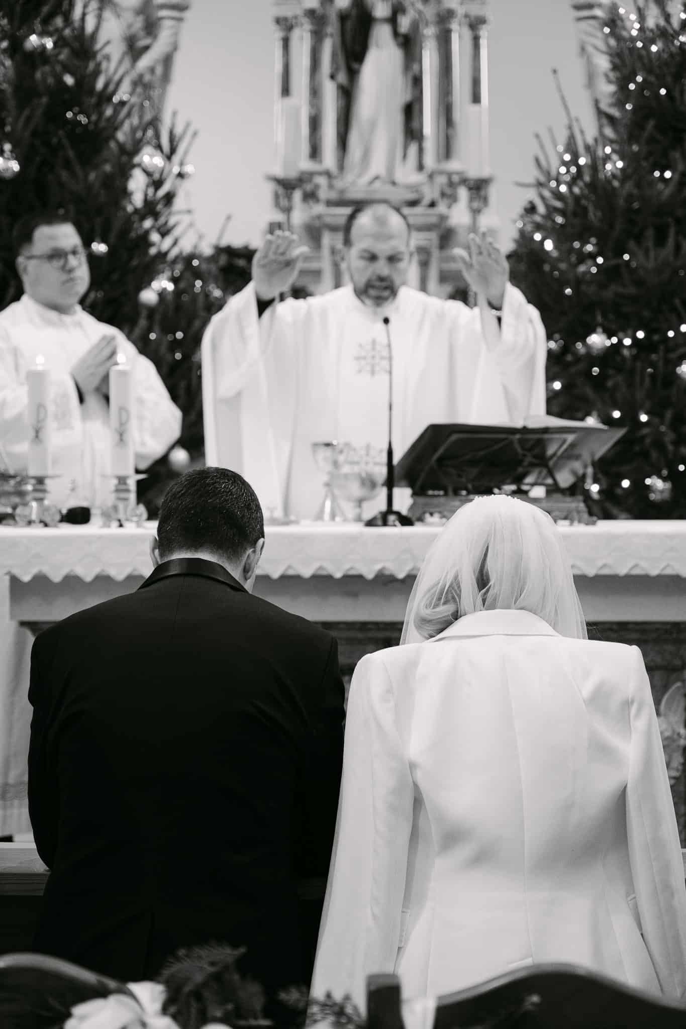 Crno-bijeli kadar crkvenog vjenčanja gdje svećenik raširenih ruku daje blagoslov mladencima koji kleče pred oltarom.