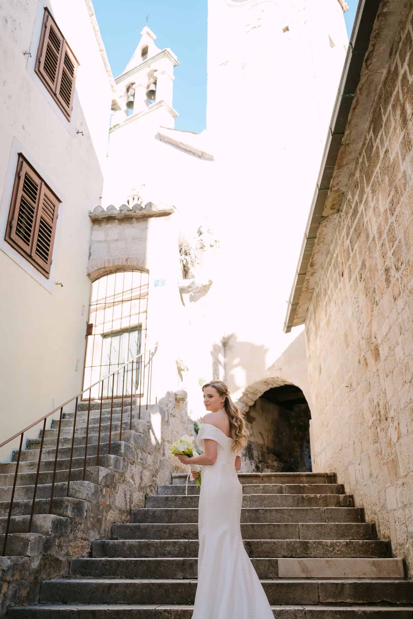 Mladenka s buketom koja pozira na starim kamenim stepenicama u dalmatinskoj ulici ispred crkvenog zvonika u gradu Omišu.