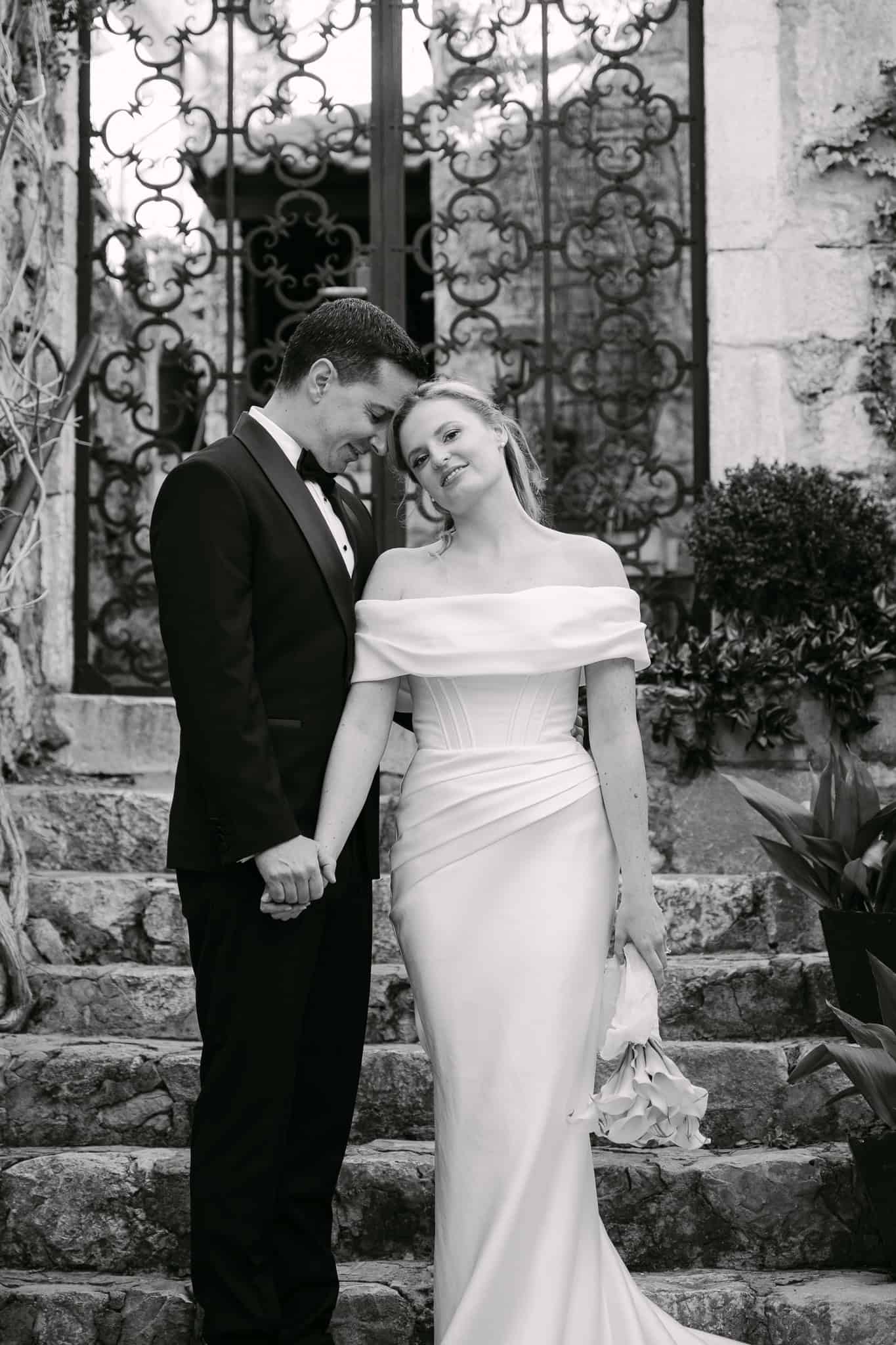 Crno-bijeli svadbeni portret mladenaca koji stoje naslonjeni jedno na drugo na kamenim stepenicama ispred vrata od kovanog željeza u centru grada Omiša.