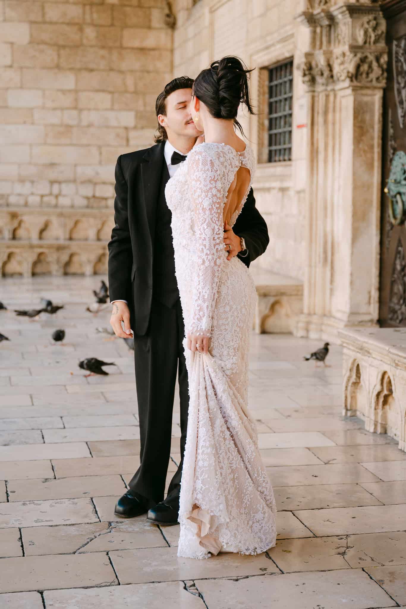 Dubrovnik wedding session na otvorenom – romantični trenutak mladenaca; mladoženja blago ljubi nevjestu u vjenčanici s otvorenim leđima ispod povijesnih kamenih arkada.
