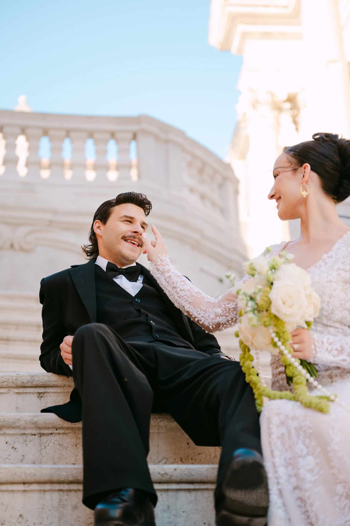 Iskren osmijeh i nježan dodir mladenaca dok opušteno sjede na suncem okupanim kamenim stepenicama; topli lifestyle trenutak s intimnog vjenčanja u povijesnom srcu Dubrovnika.