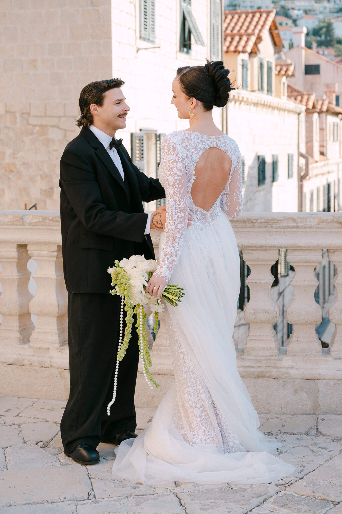 Nježni susret mladenaca na kamenoj terasi; mladenka u vjenčanici s otvorenim leđima i raskošnim buketom gleda mladoženju ispred autentičnih dubrovačkih kuća i brda u daljini.