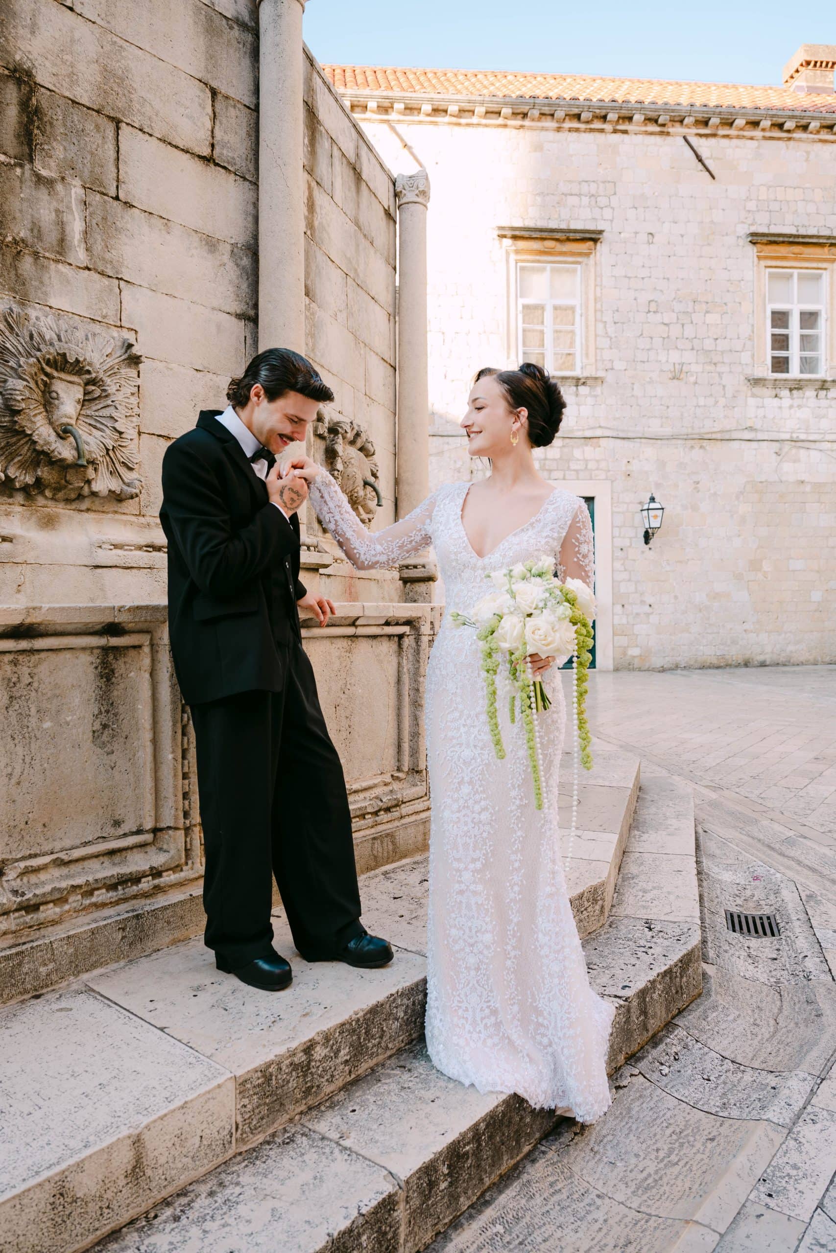 Dubrovnik - romantičan vjenčani trenutak na skalinama; mladoženja u crnom odijelu ljubi ruku mladenki ispred starog kamenog zida u ljetno popodne.