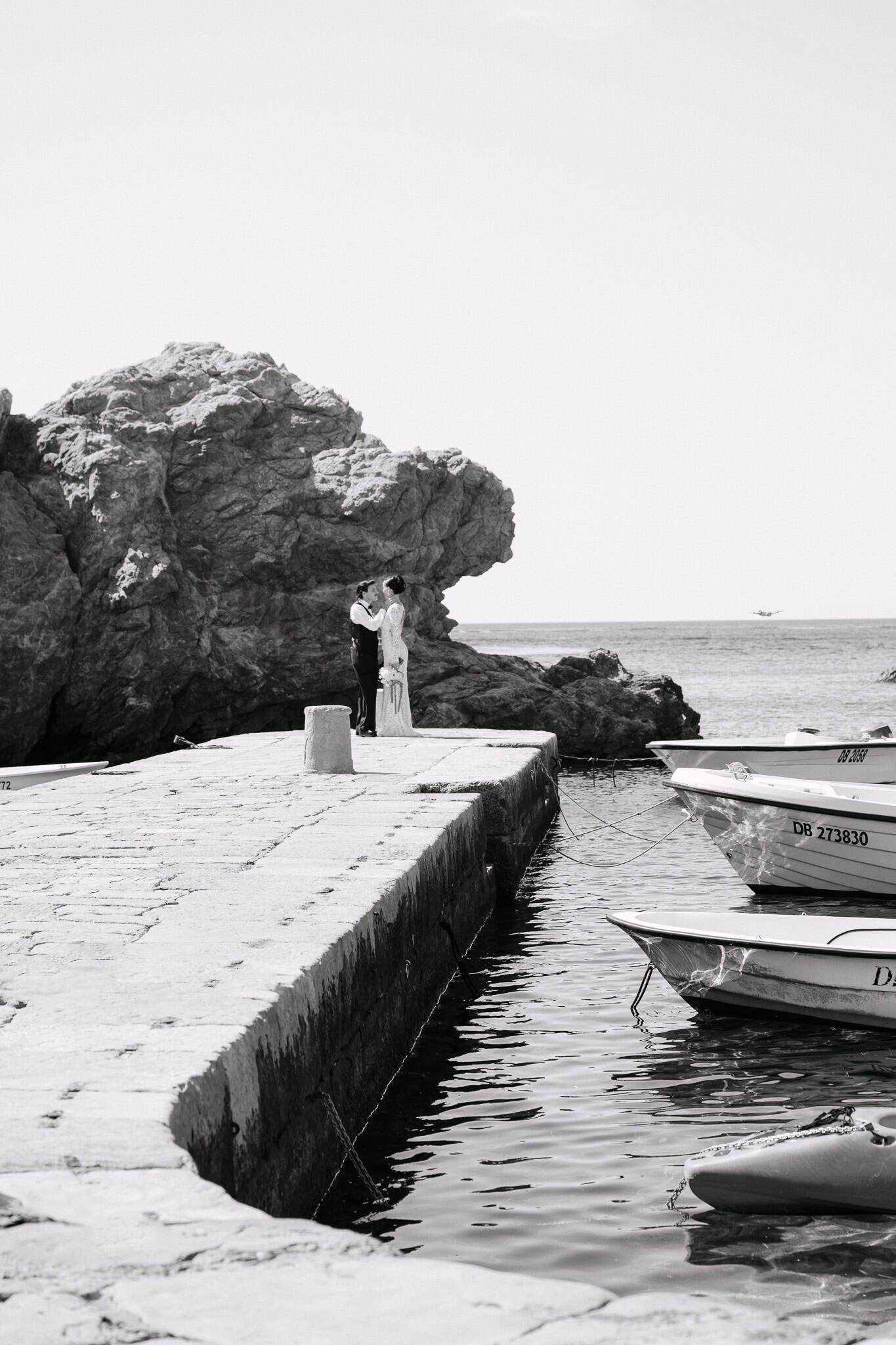 Vjenčanje u Dubrovniku - romantična crno-bijela fotografija uz more; mladenci poziraju na kamenom pristaništu ispred impresivnih morskih hridi i usidrenih čamaca.