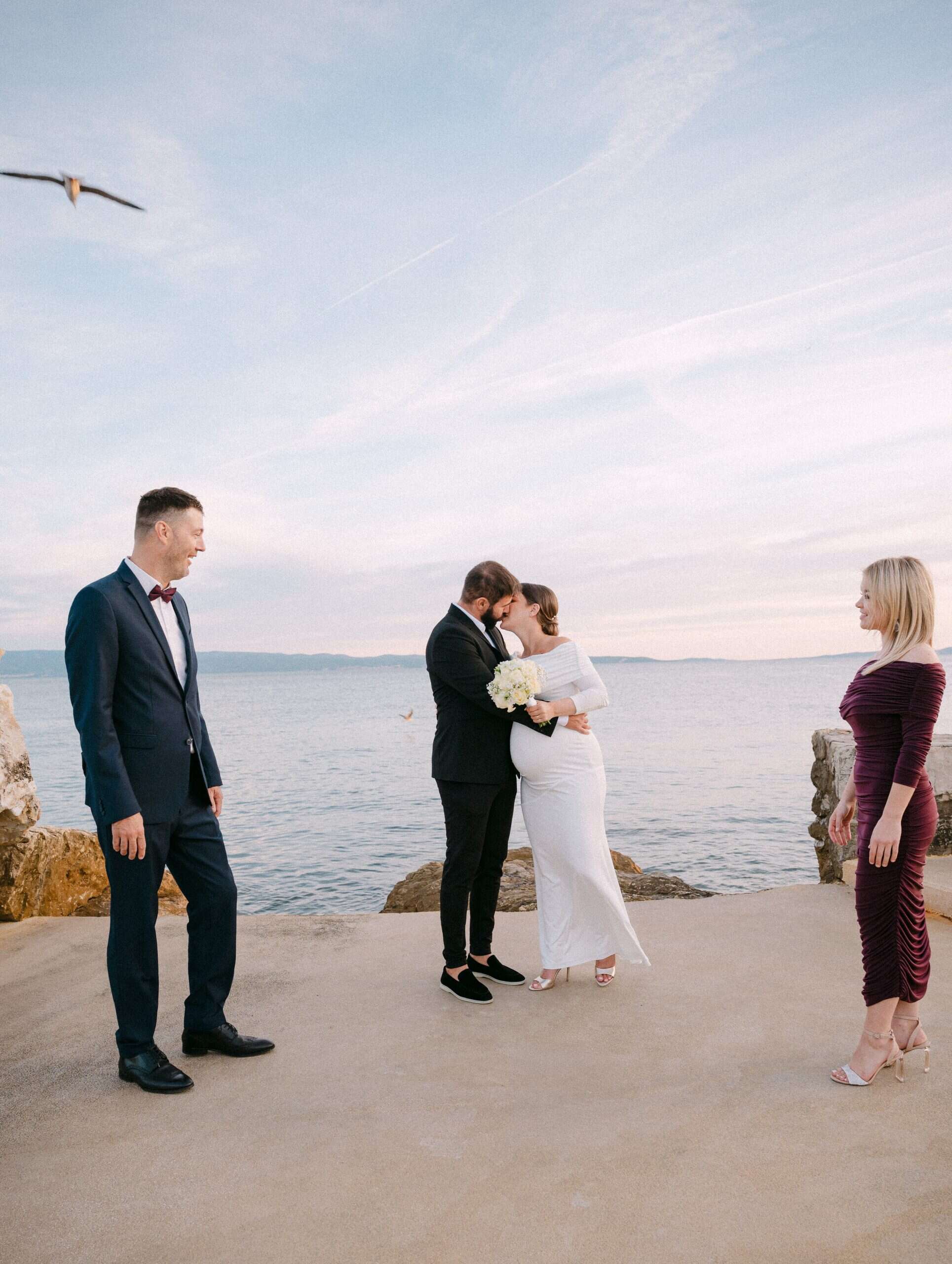 Fotografija s vjenčanja, mladenci i kumovi, koja prikazuje ljubav i radost uz more u zalazak sunca.