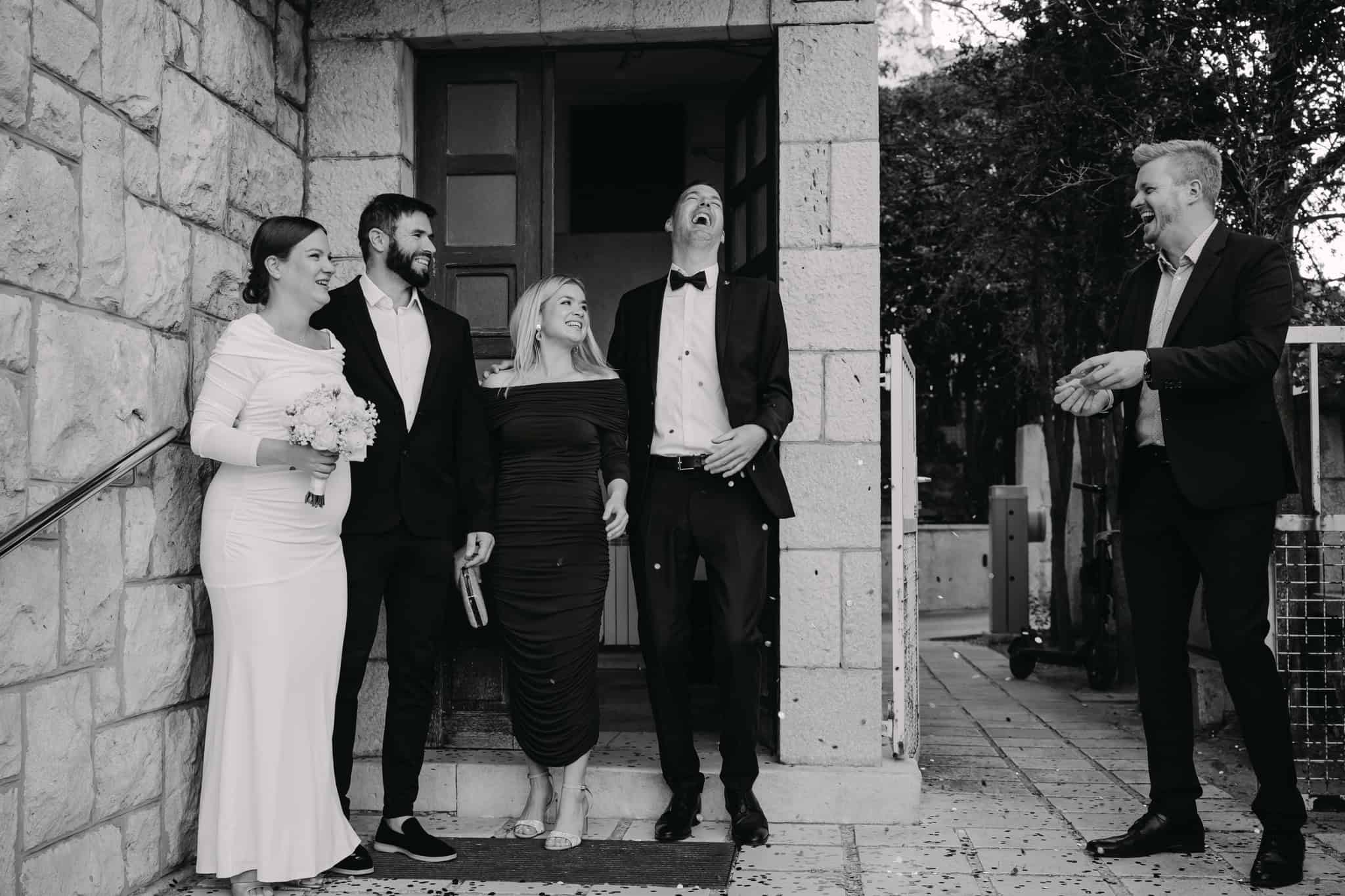 Upravo vjenčani mladenci i prijatelji veselo se smiju ispred kamene zgrade.