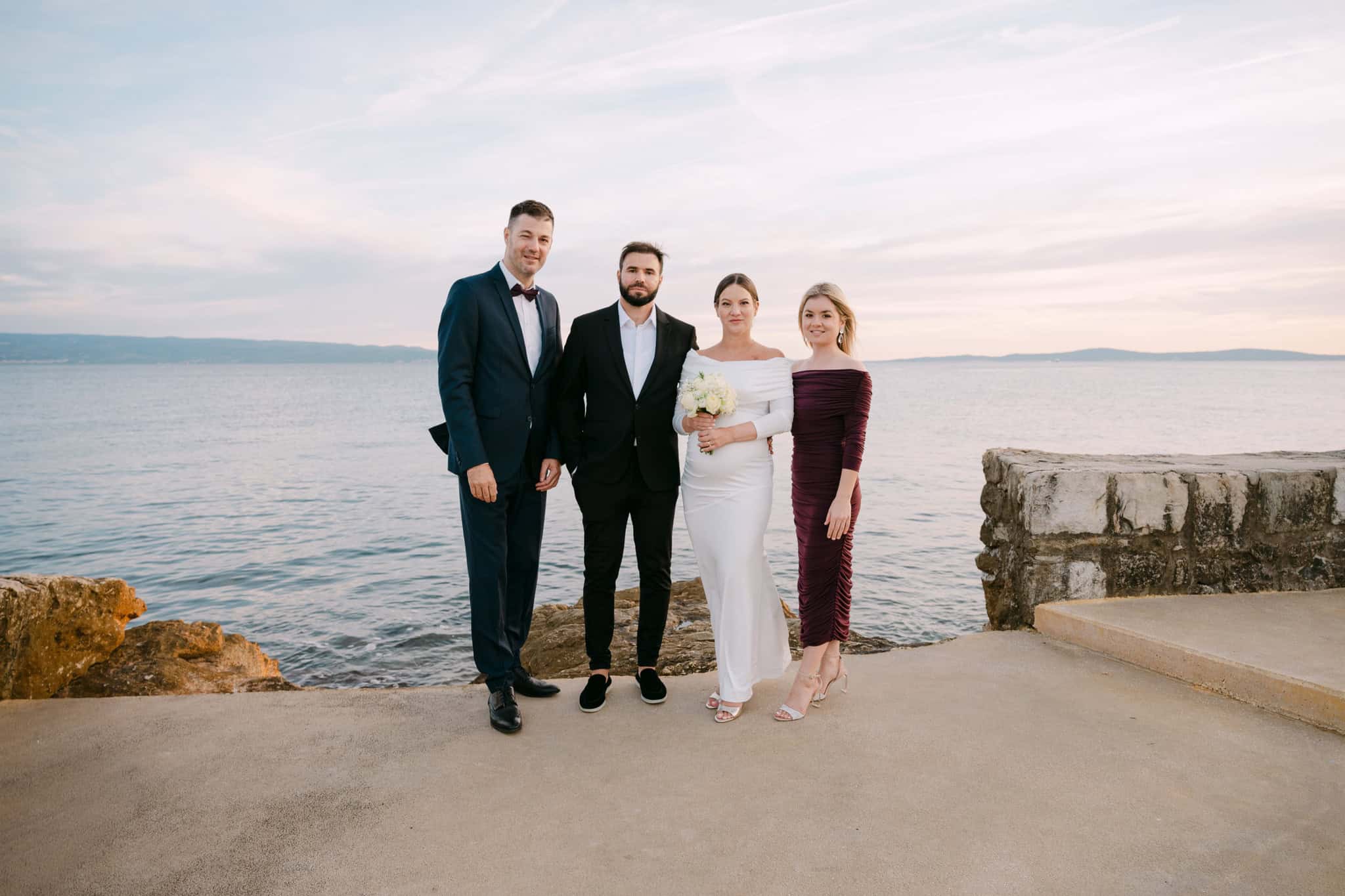 Svadbeni par s kumovima stoji uz obalu tijekom zalaska sunca, odjeveni u svečanu odjeću s pogledom na Jadransko more.