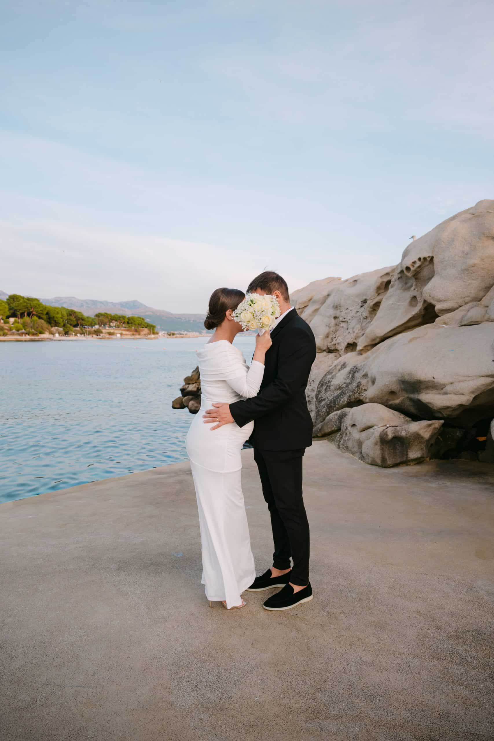 Portret mladenaca na Bačvicama u Splitu; trudna mladenka u bijeloj haljini drži buket ispred lica mladenca, velika kamena stijena i more u pozadini.