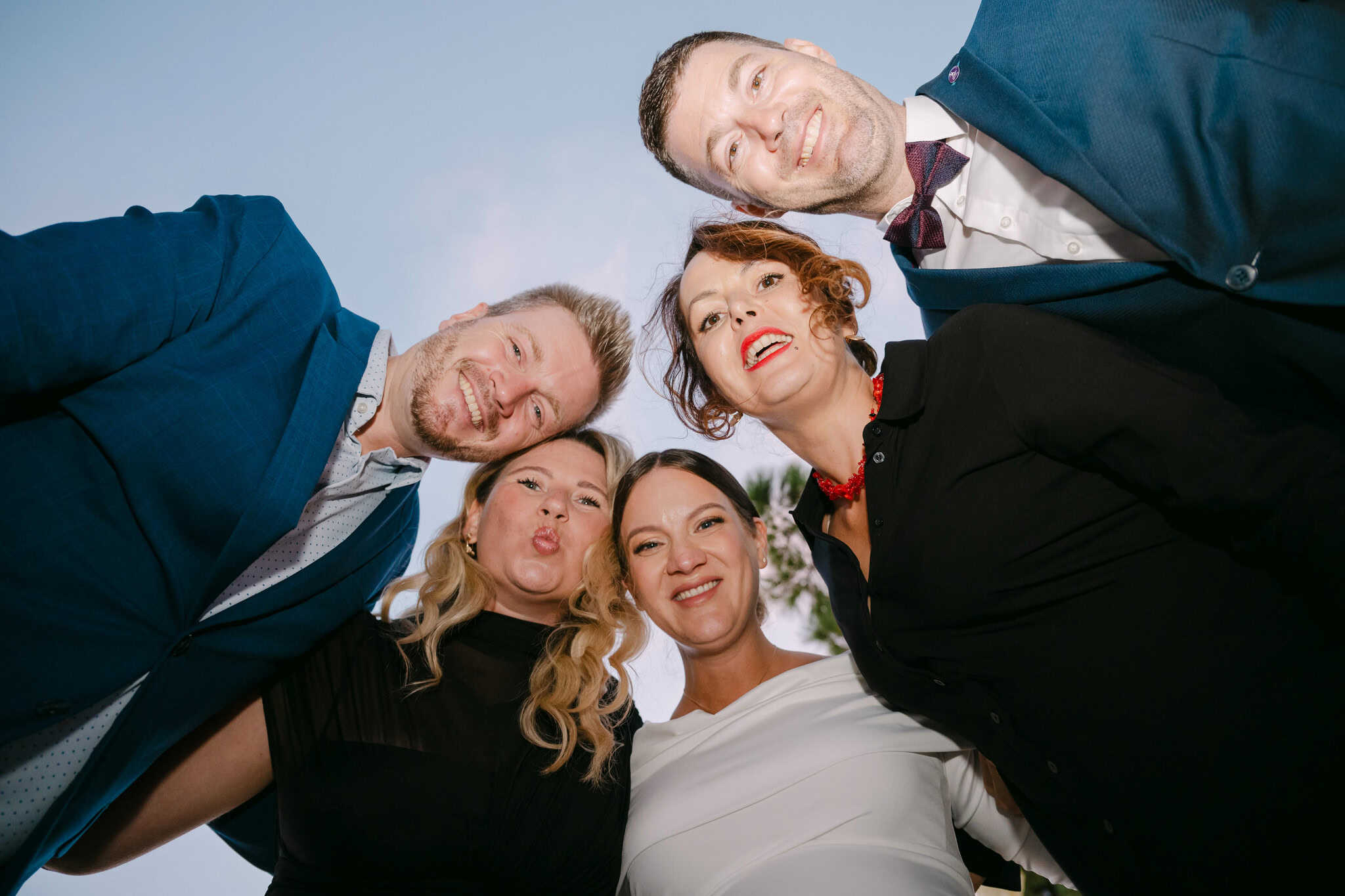 Zabavan grupni portret mladenke i gostiju slikan iz donjeg kuta; nasmijana lica u krugu gledaju prema kameri na vjenčanju u Splitu.