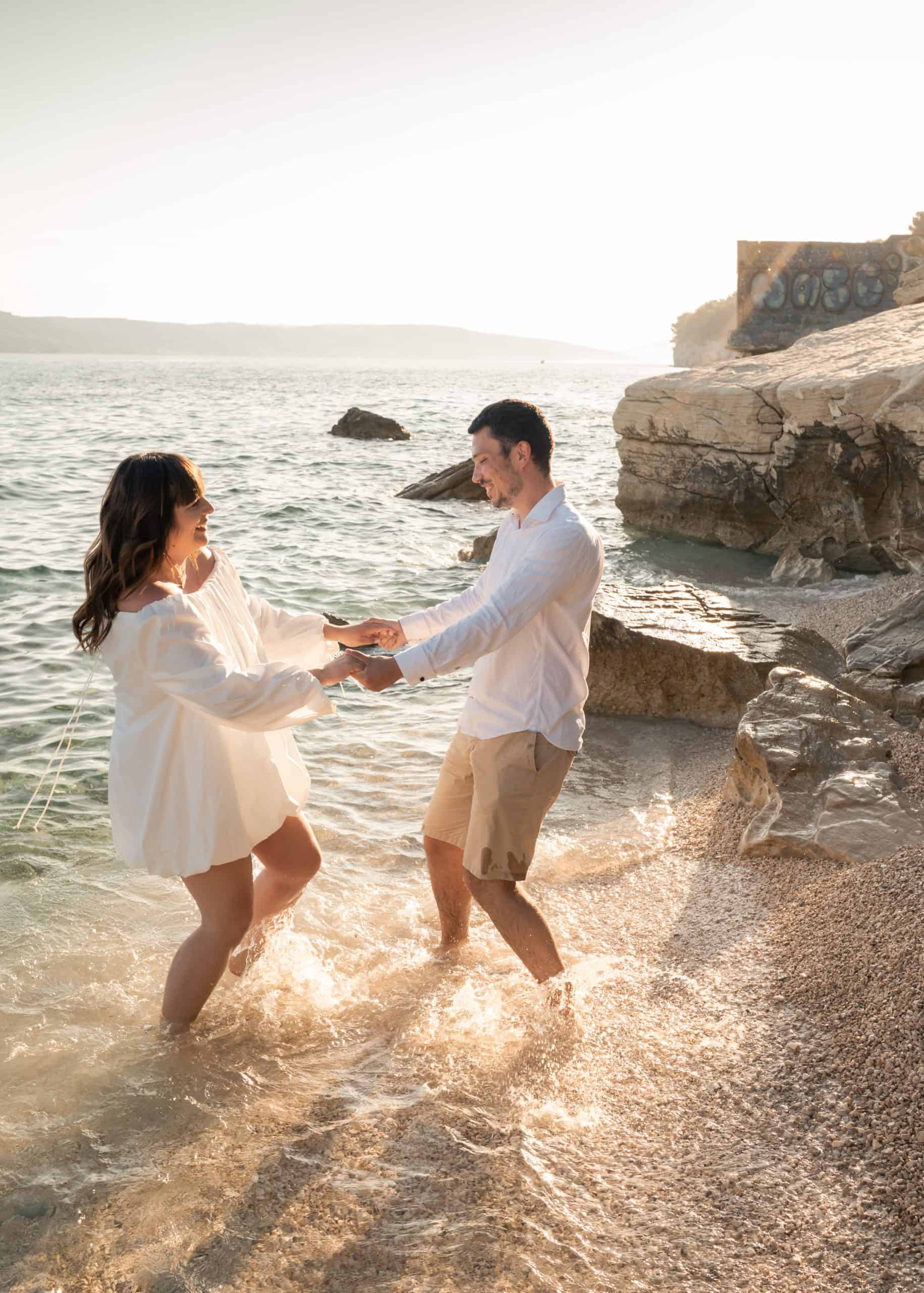 Ljubavni par na plaži, držeći se za ruke i uživajući u romantičnoj atmosferi uz more i stijene.
