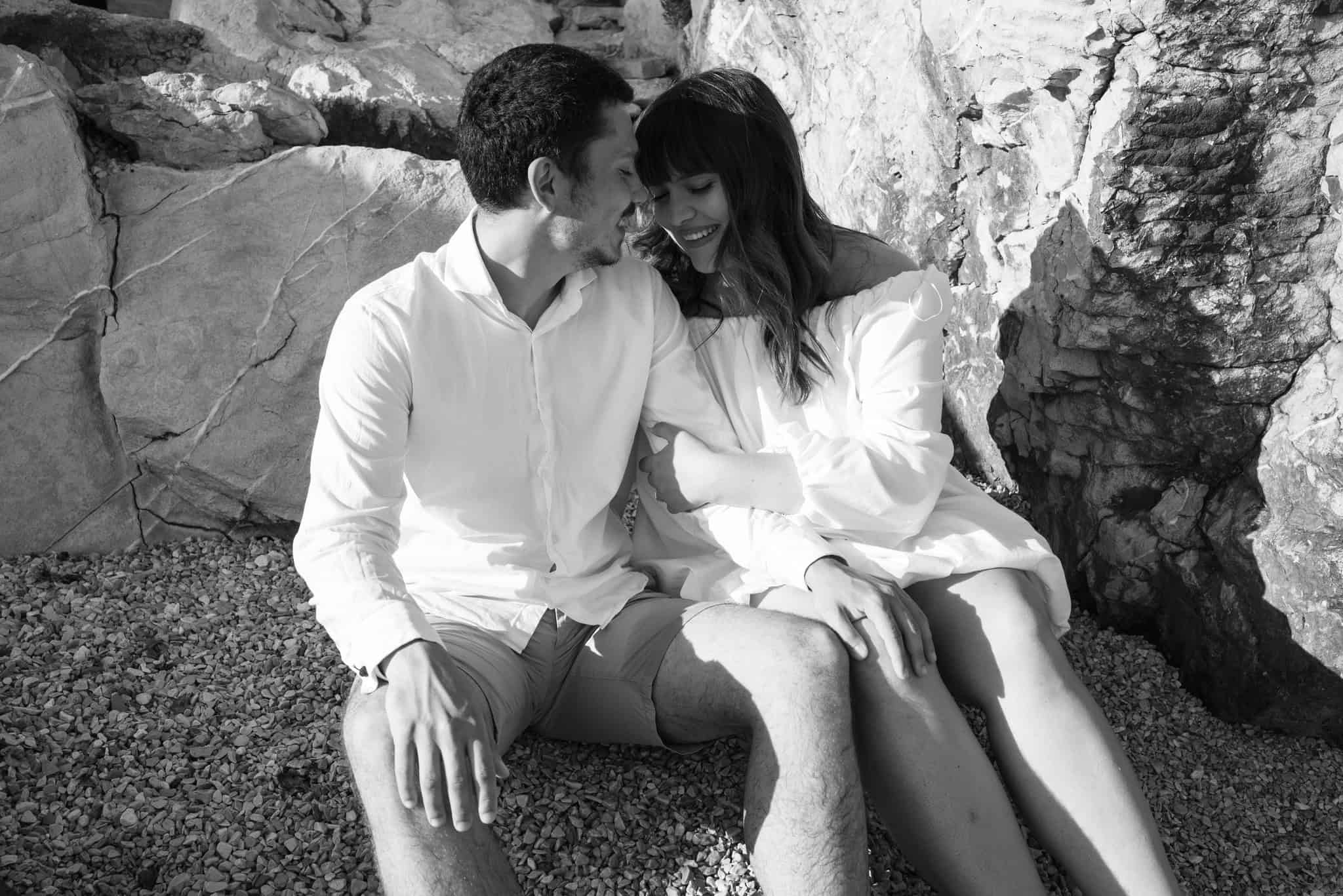 Zaljubljeni par naslonjen jedno na drugo i smiješi na plaži s kamenjem u pozadini.