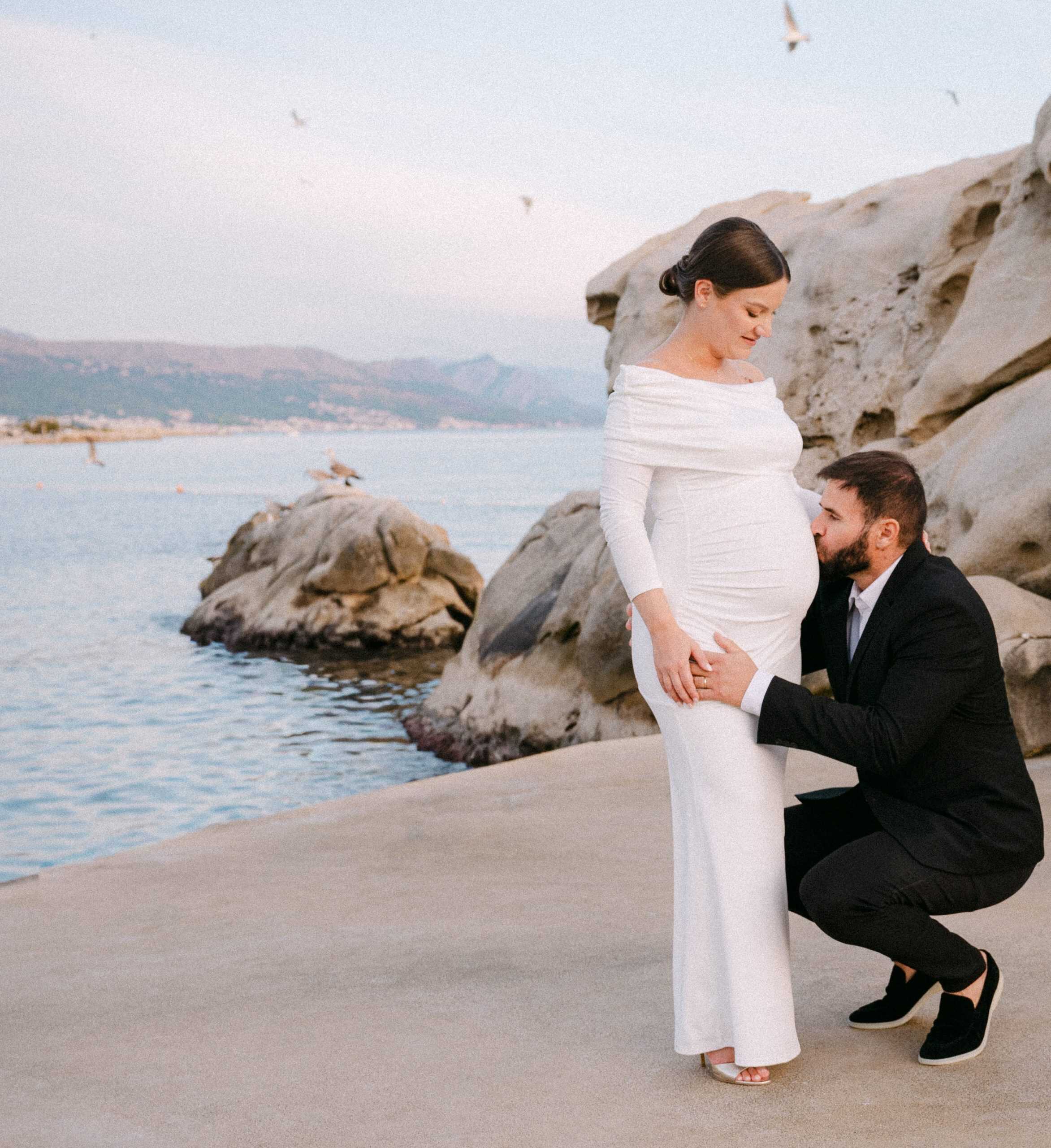 Trudnica u bijeloj haljini s partnerom koji kleči pokraj nje na stjenovitoj obali.